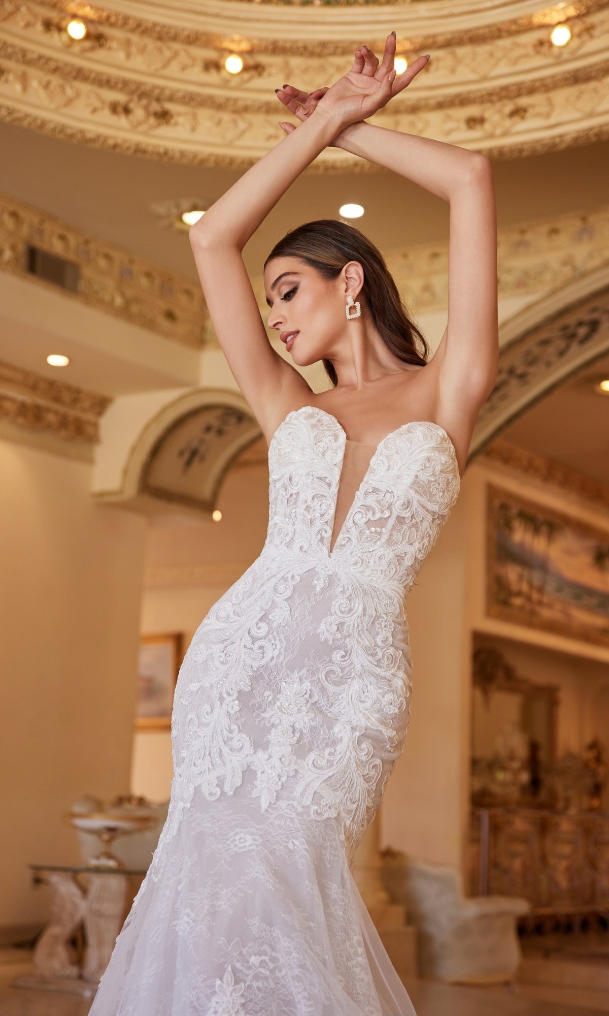A woman poses with her arms raised in an ornate, elegant interior while wearing the Ladivine CD928 Strapless Mermaid Wedding Gown featuring lace and a deep neckline.