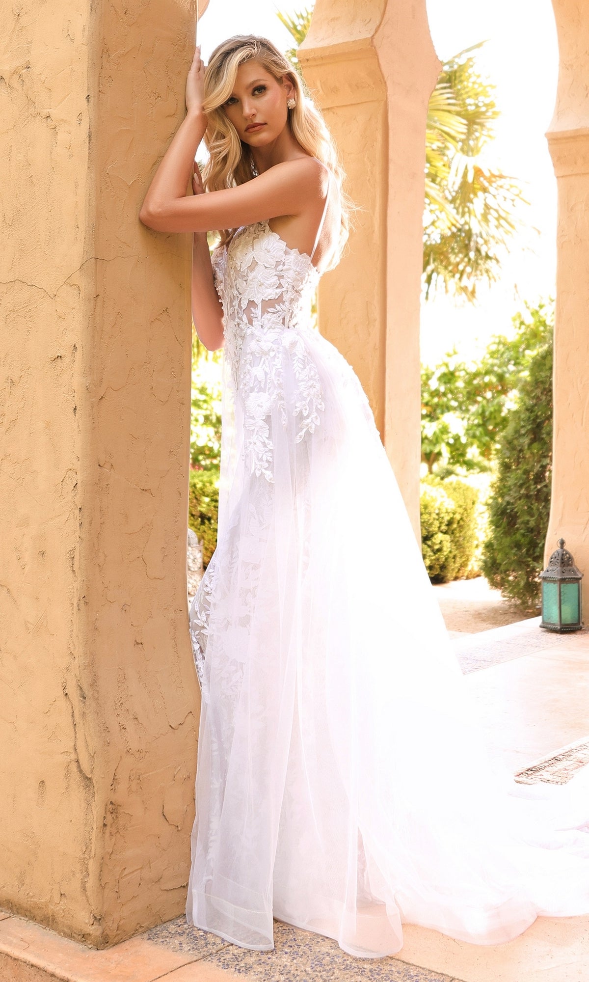 A woman wearing the Sheer-Corset Long White Lace Wedding Dress CD931W with a sheer overskirt stands by a tan stucco pillar outdoors, surrounded by greenery and sunlight.