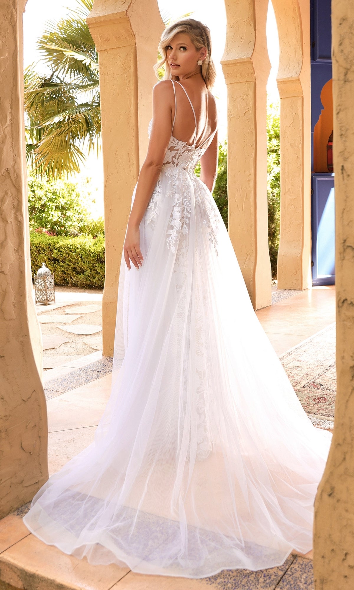 A woman in the Sheer-Corset Long White Lace Wedding Dress CD931W with a long sheer overskirt stands outdoors among architectural columns, looking over her shoulder.