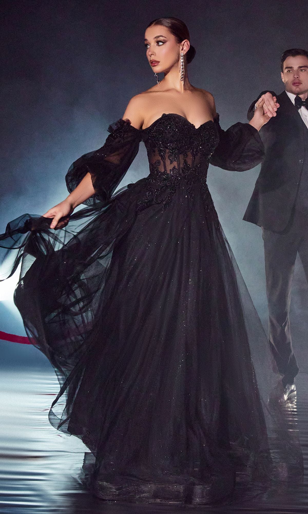 A woman wears the Strapless Puff-Sleeve Long Prom Dress CD948, featuring removable puff sleeves and intricate detailing, as she stands on a reflective floor holding hands with a man in a black tuxedo.