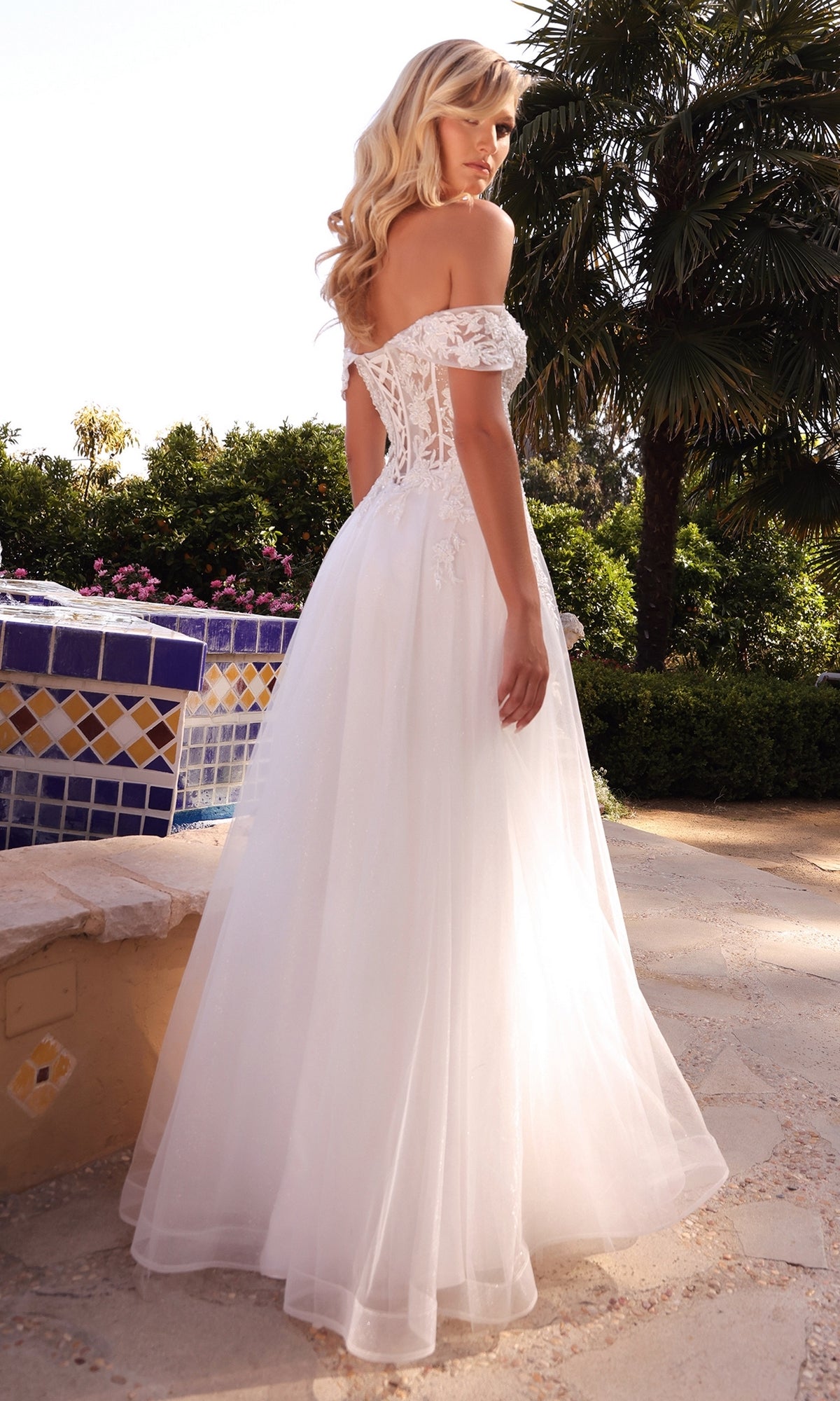 A woman in the Sheer-Bodice Long White Prom Ball Gown CD961W, featuring an off-the-shoulder lace bodice and lace-up back, stands outdoors by a tiled fountain amid lush greenery.