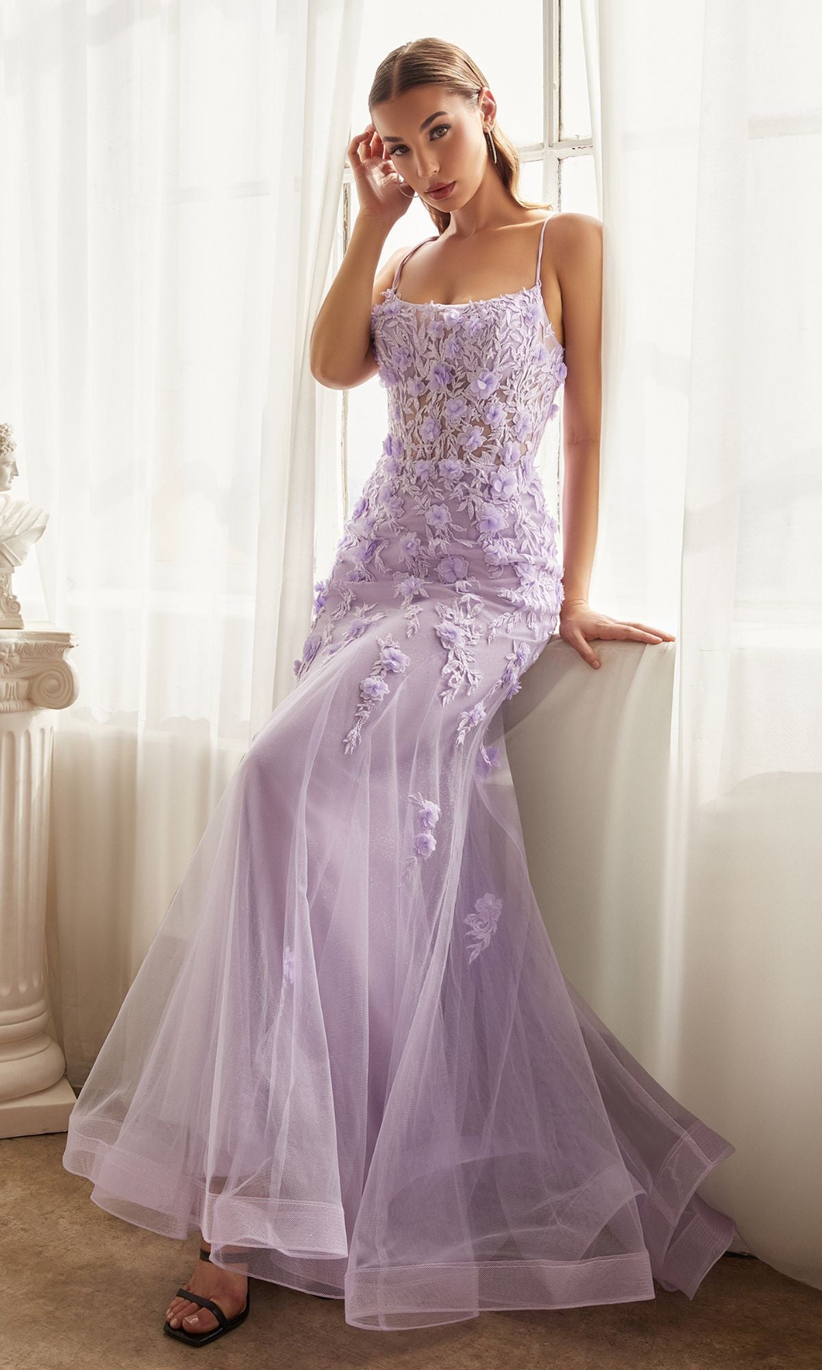 A woman stands indoors by a window with white curtains, wearing the Floral-Applique Long Mermaid Prom Dress CD995 in lavender, featuring sheer fabric and floral embroidery.