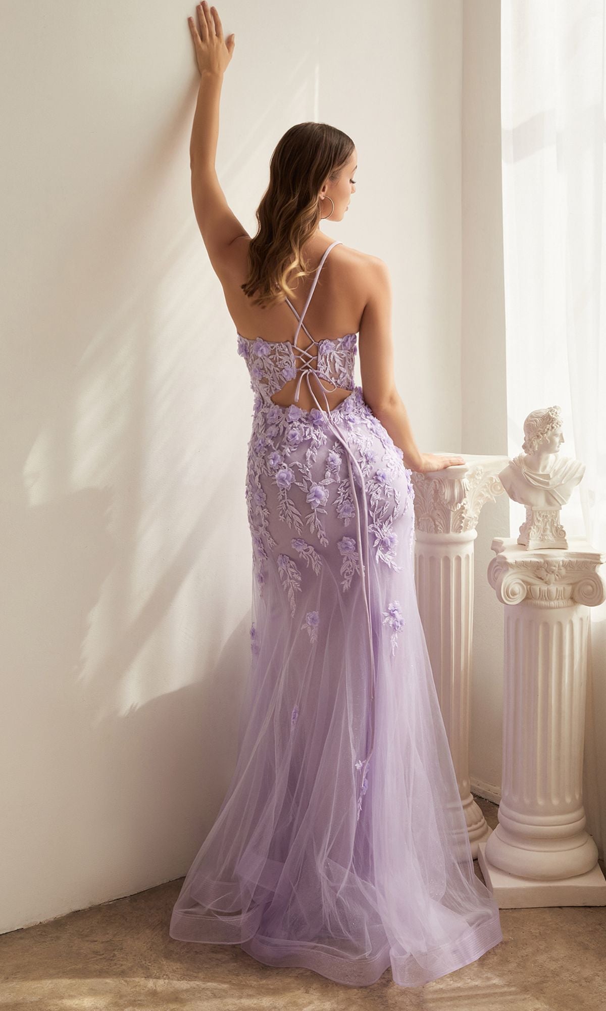 A woman wearing the Floral-Applique Long Mermaid Prom Dress CD995 stands with her back to the camera, one arm raised, beside white columns and sheer curtains in a softly lit room.