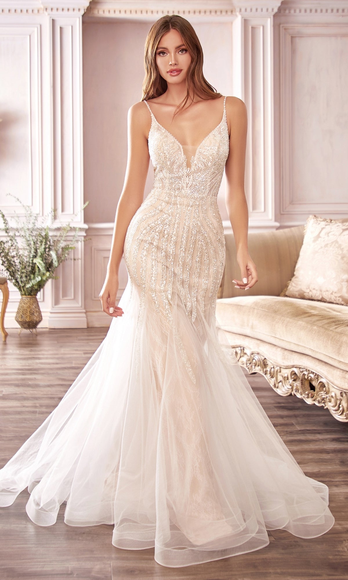 A woman in the Off-White Long Beaded Mermaid Formal Dress CDS401, featuring a fitted sleeveless design and sheer tulle skirt, stands in an elegant room with ornate decor and a beige sofa.