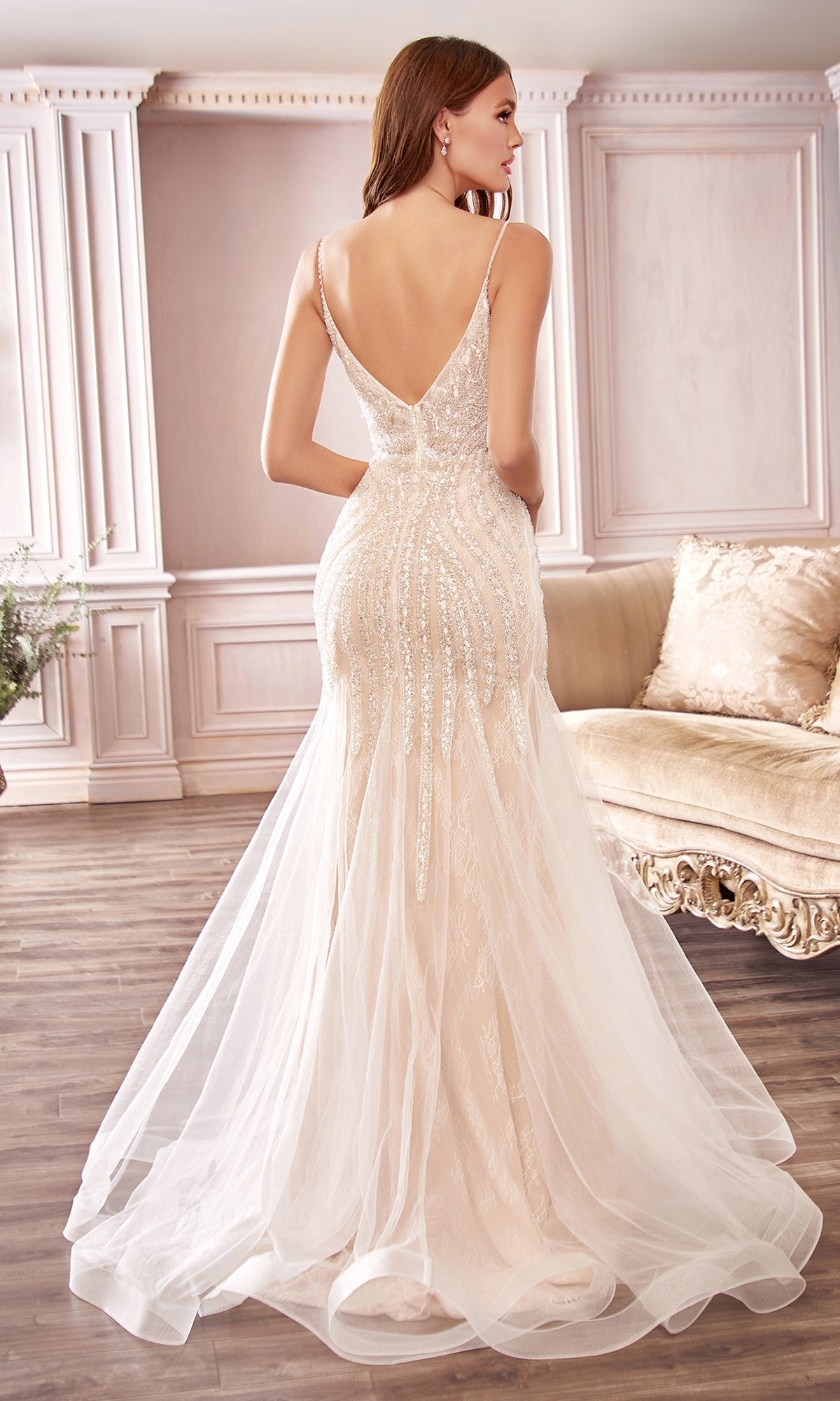 A woman stands in an elegant room with wooden floors and classic decor, wearing the Off-White Long Beaded Mermaid Formal Dress CDS401—a fitted, floor-length gown featuring white lace, a deep V-back, and beaded detailing.