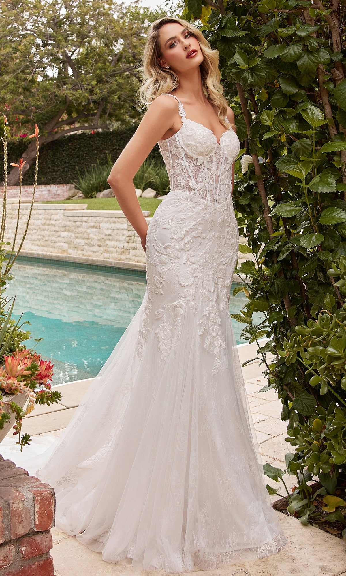 A woman stands by a pool amid lush greenery and brick accents, wearing the Chantilly-Lace Long Off-White Formal Dress CDS432W featuring elegant lace details and a corset-style waist.