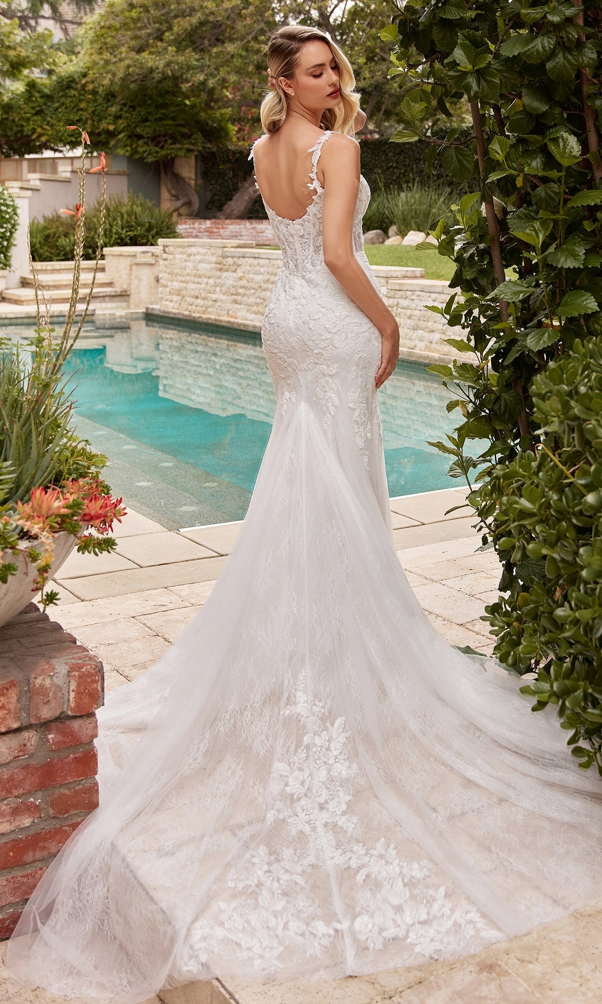 Wearing the Chantilly-Lace Long Off-White Formal Dress CDS432W, a woman stands by a pool in a beautiful garden, showcasing the gown's corset-style waist and long train as she faces away from the camera.