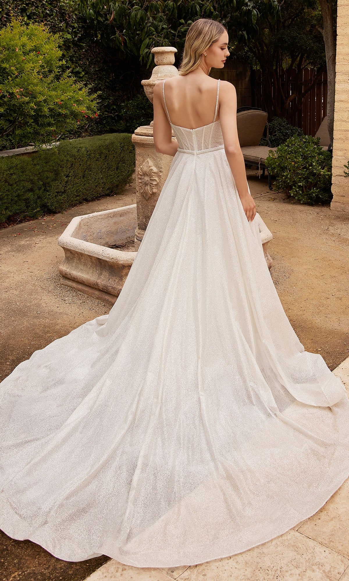 A woman in the Off-White Bustier-Bodice Long Formal Dress CDS435W, featuring a sweeping train, stands outdoors near a stone fountain, facing away.