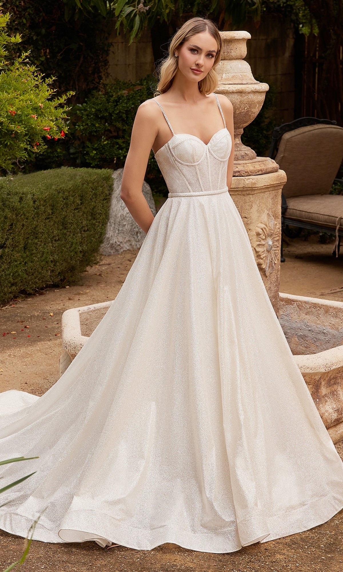 A woman stands outdoors in the Off-White Bustier-Bodice Long Formal Dress CDS435W—a sleeveless, floor-length gown with a fitted bodice and thin straps. Bushes, a stone bench, and a fountain add elegance to the scene.