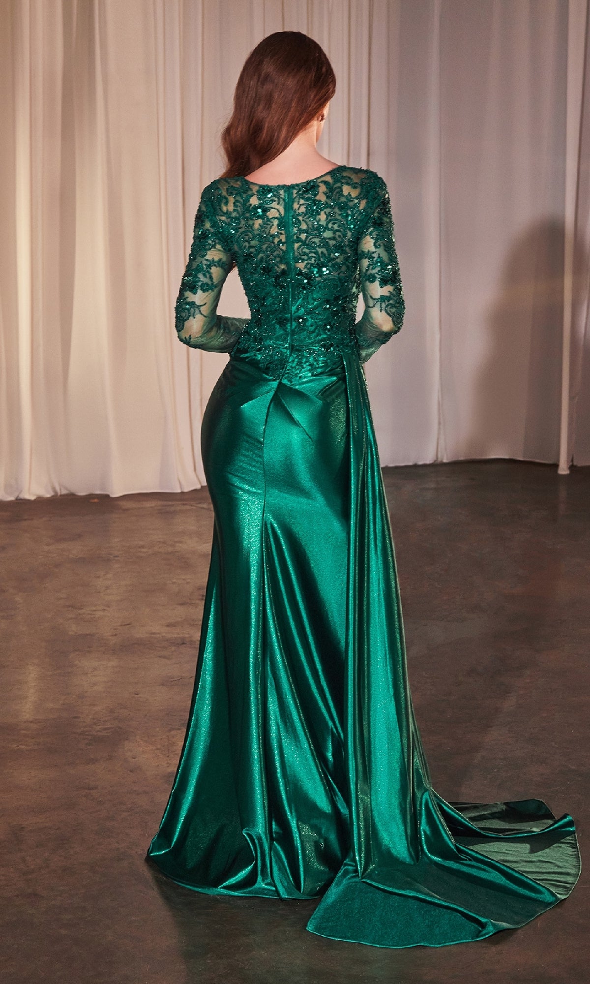 A woman in the Ladivine CDS506, a floor-length emerald green formal dress with lace-detailed long sleeves and upper back, stands facing away in a dimly lit room with white curtains.