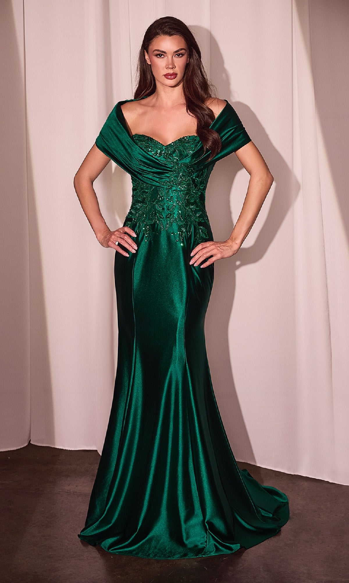 A woman poses by a white curtain in the Ladivine CDS507, an emerald green off-shoulder wrap long prom dress with a sweetheart neckline, satin fabric, and lace with bead detailing on the bodice.