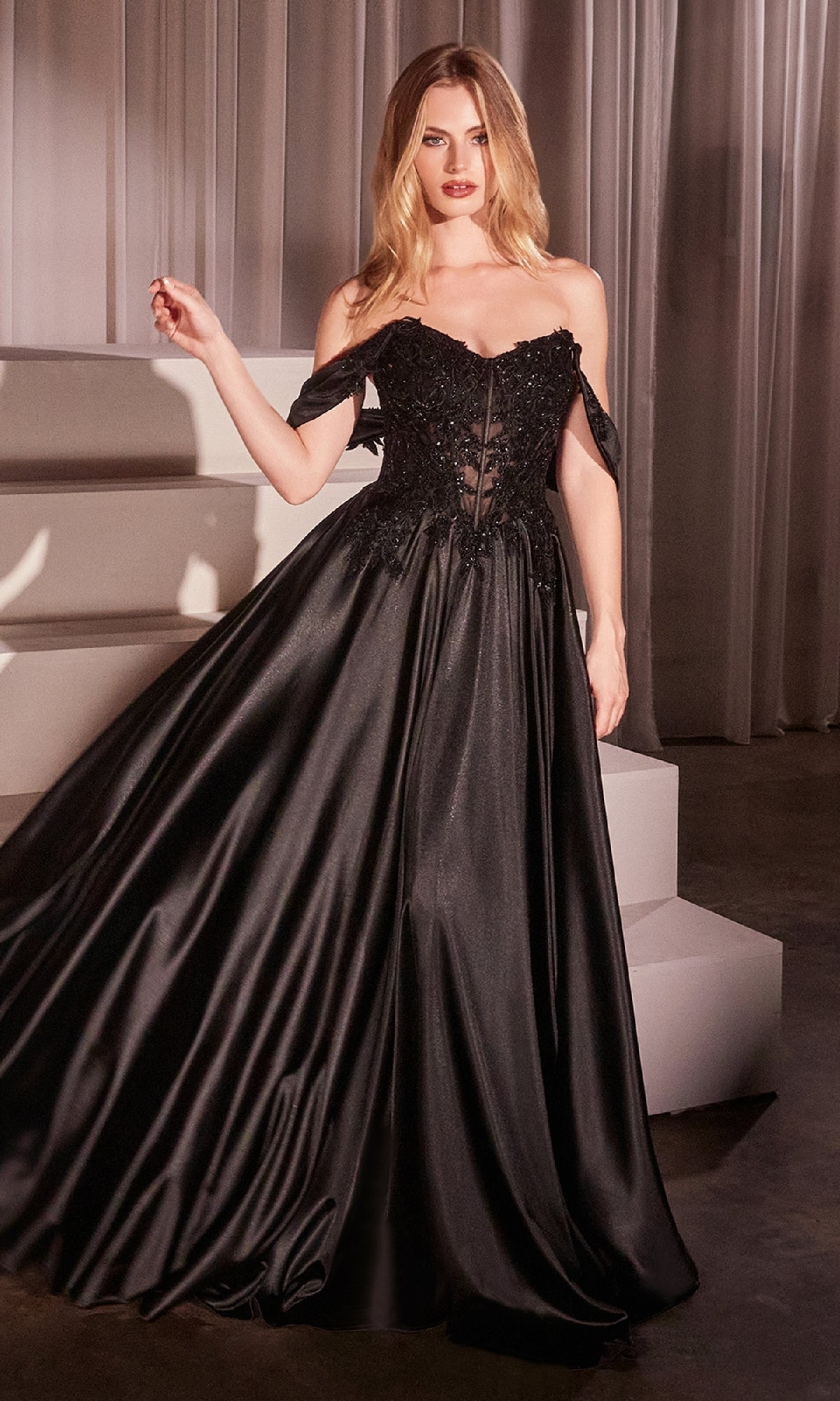 A woman in the Off-the-Shoulder Prom Ball Gown: Ladivine CDS525, a black evening dress with a flowing skirt and sheer bodice, stands indoors by white steps and draped curtains.