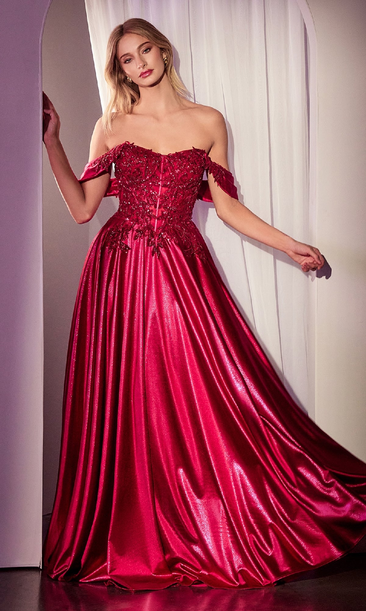 A woman stands in a doorway wearing the Off-the-Shoulder Prom Ball Gown: Ladivine CDS525, a shiny red formal dress with floral embellishments on the bodice and a graceful, floor-length silhouette.