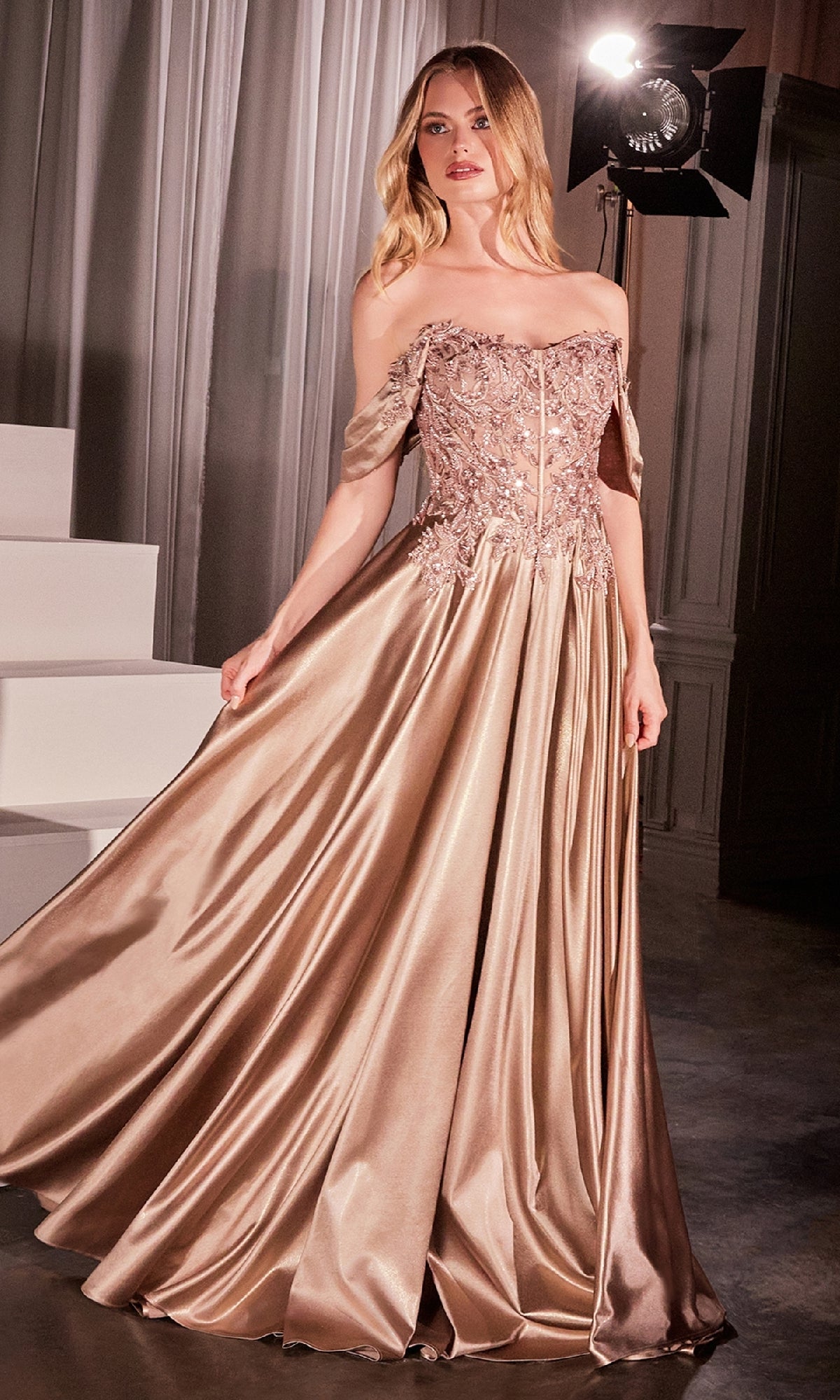 A woman stands indoors by a staircase in soft light, wearing the Off-the-Shoulder Prom Ball Gown: Ladivine CDS525—an elegant gold satin dress with floral embellishments.