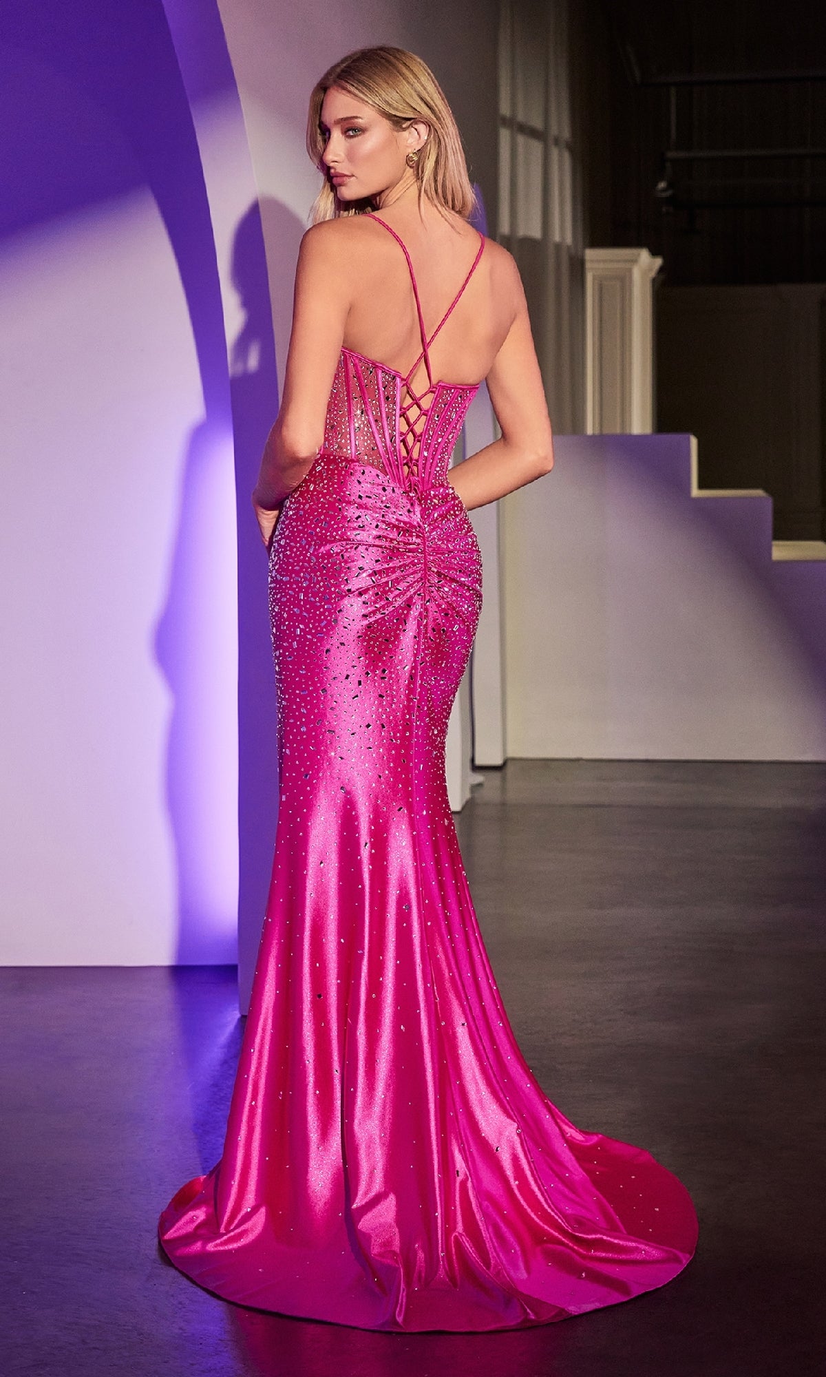 A woman in the Sheer-Corset Beaded Prom Gown: Ladivine CDS527, a fitted floor-length bright pink dress with sparkling embellishments and a lace-up back, stands facing away indoors.
