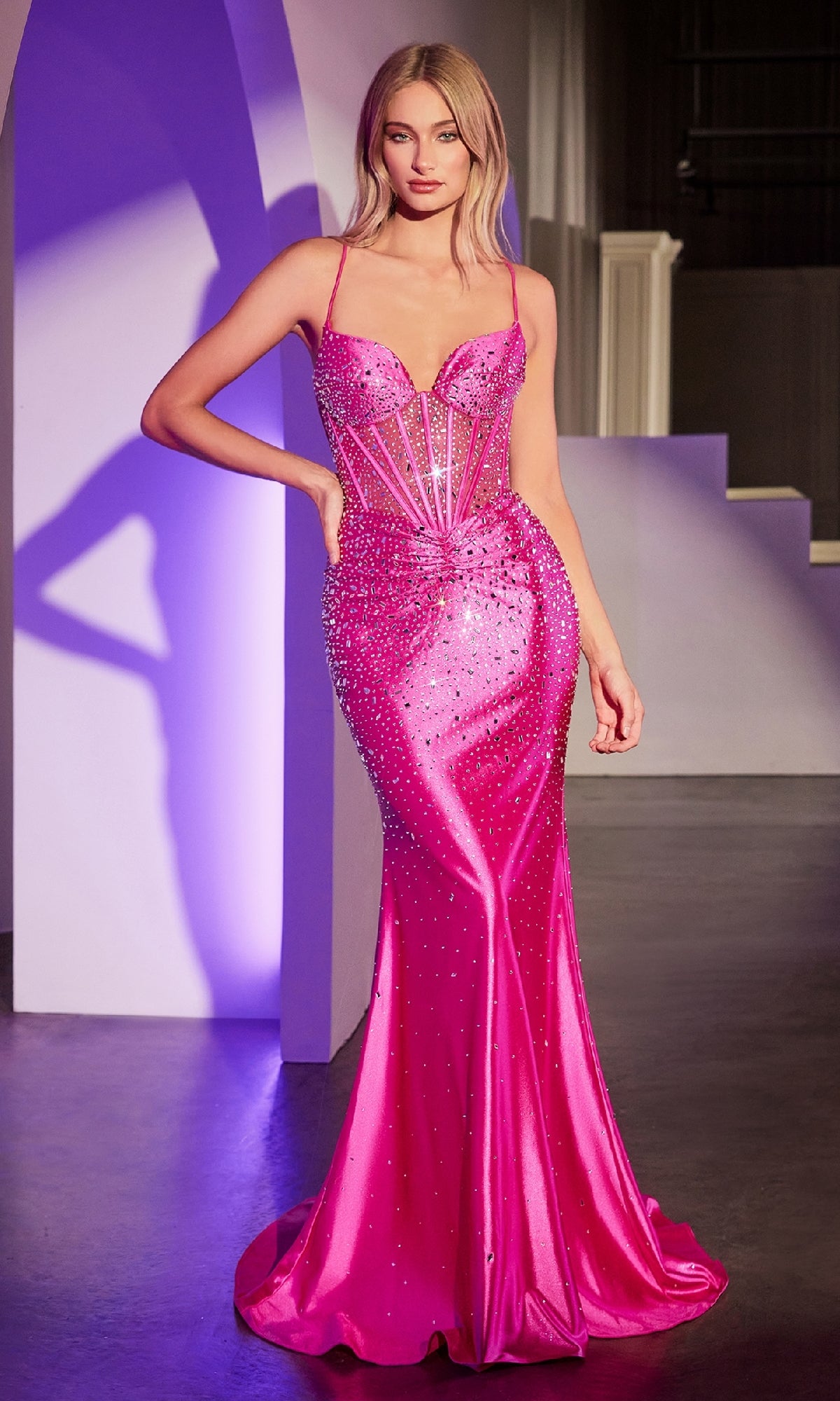A woman stands indoors in the Sheer-Corset Beaded Prom Gown: Ladivine CDS527, a bright pink, floor-length dress with thin straps, delicate embellishments, and a sheer corset bodice—ideal for proms or special occasions.