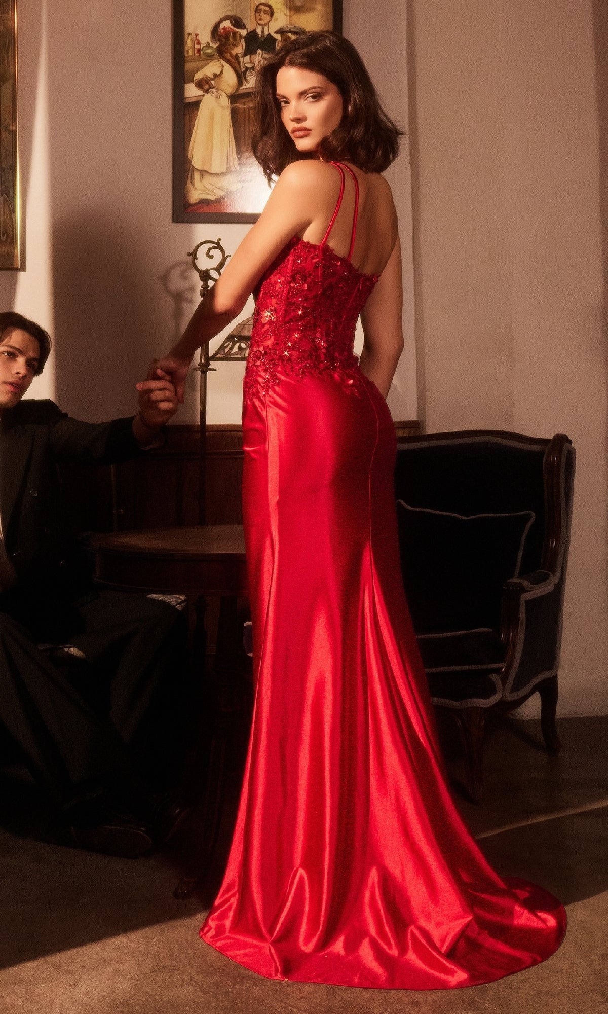 A woman in the Ladivine CDS535 One-Shoulder Long Prom Dress stands indoors, facing away but looking over her shoulder, as a seated person holds her hand. Paintings are displayed on the wall behind them.