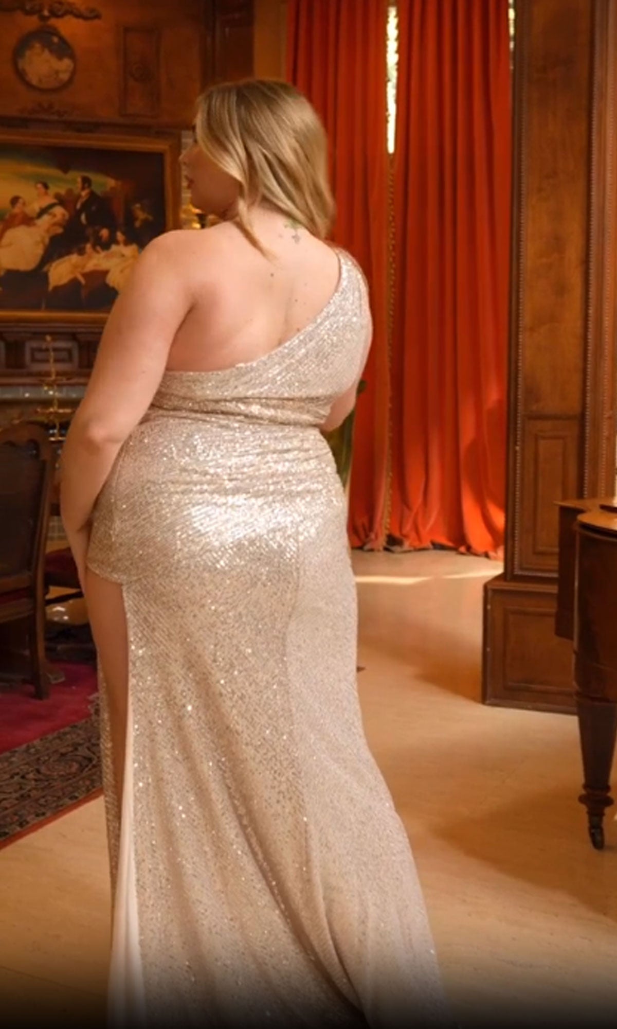 A person in the One-Shoulder Plus-Size Sequin Prom Dress CH077C stands indoors, facing away in an elegant wood-paneled room with red curtains. The floor-length sequin gown sparkles, adding glamour to the sophisticated setting.