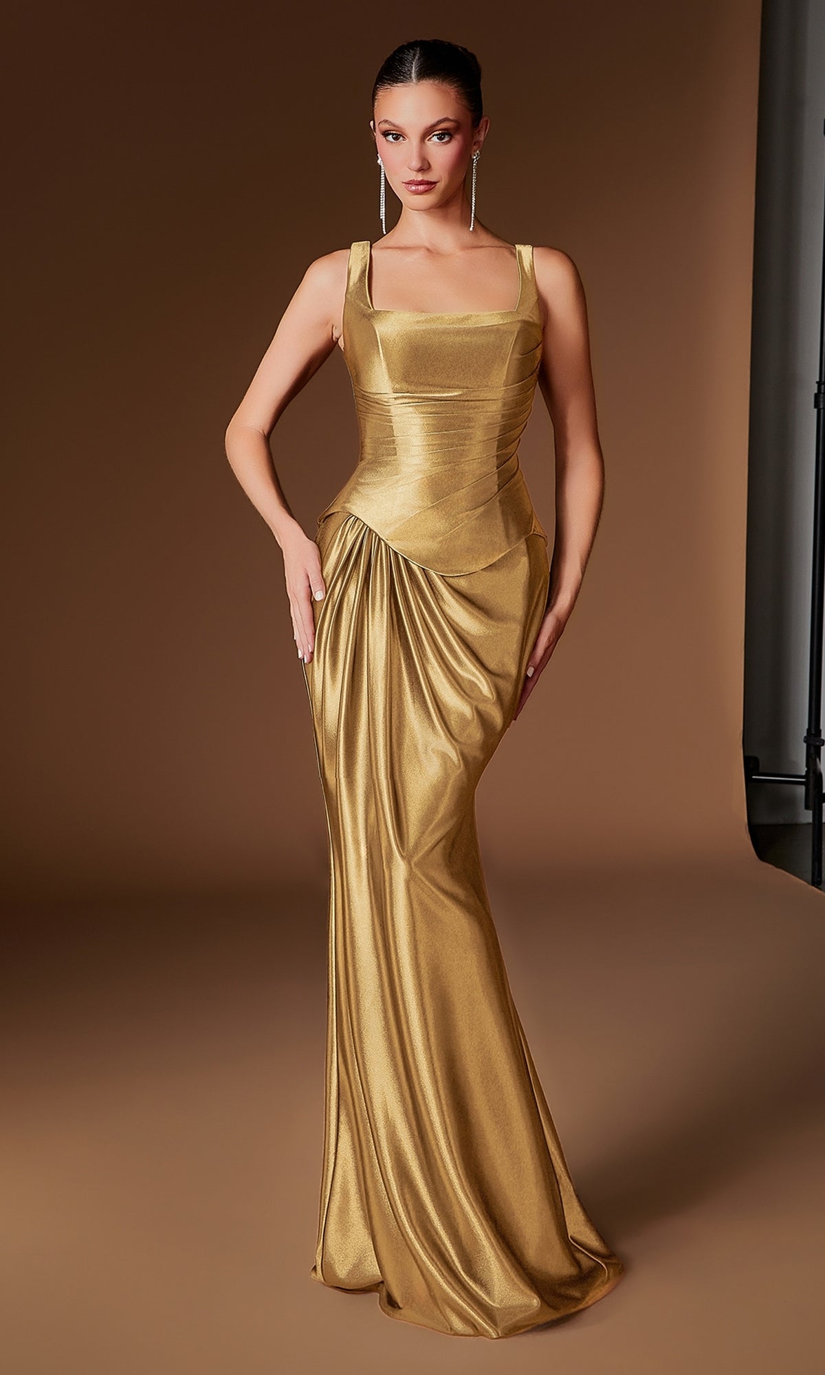 A woman wearing the Ladivine CH2604 long prom dress in gold, which features a form-fitting design with a pleated satin bodice, draped skirt, structured silhouette, and elegant square neckline, stands against a plain brown background.