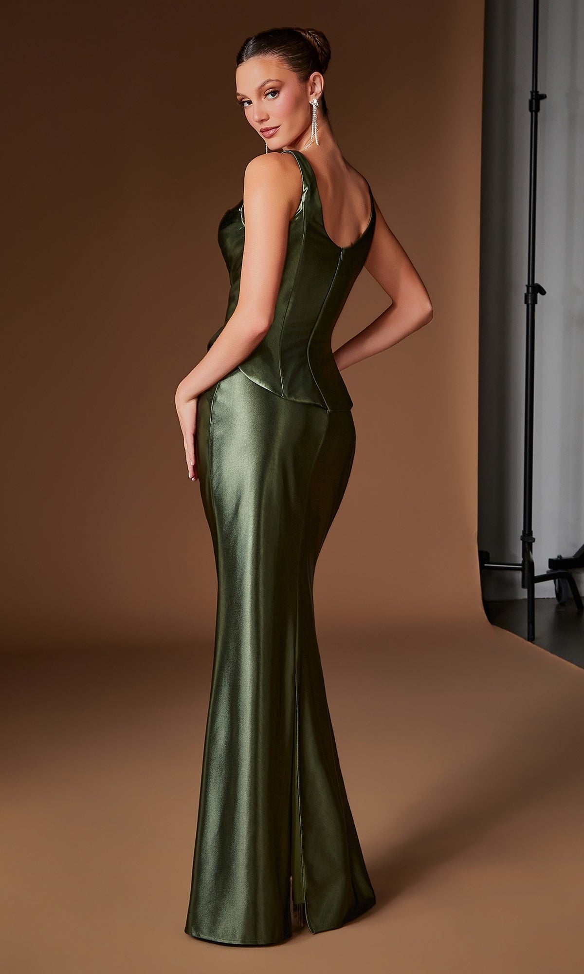 A woman wearing the Ladivine CH2604 long prom dress—featuring a fitted, metallic olive green silhouette with a pleated satin bodice—stands on a brown backdrop, facing slightly away and glancing over her shoulder.