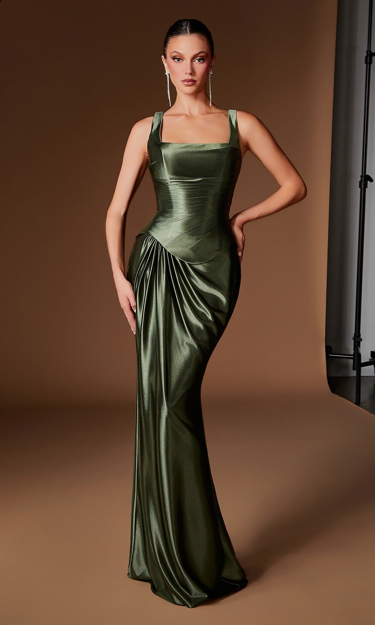 A woman stands against a plain brown studio backdrop wearing the Ladivine CH2604 long prom dress in olive—a shiny, fitted gown featuring a pleated satin bodice, square neckline, shoulder straps, and elegant draped detailing.
