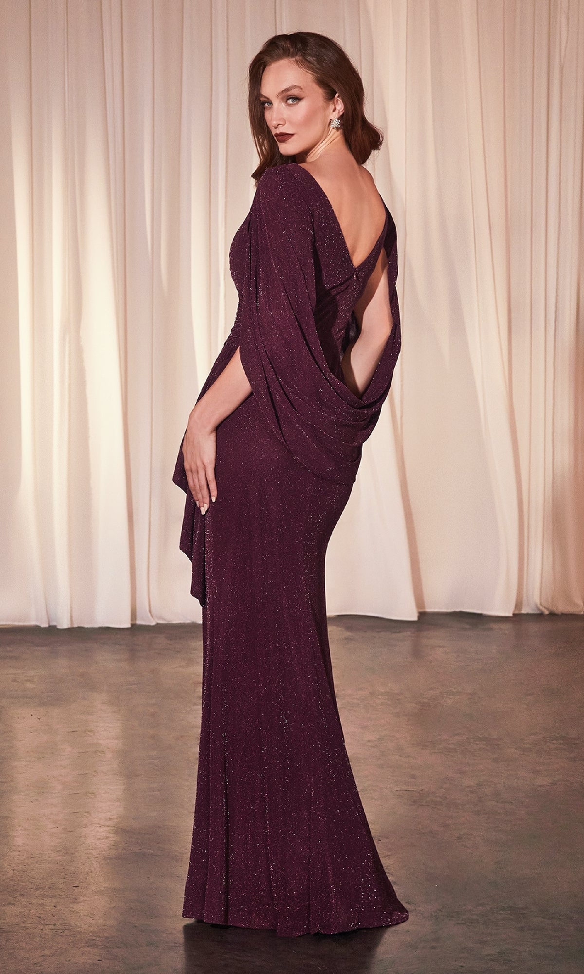 A woman wearing the Ladivine CH386 Wine Red Long Wedding-Guest Dress with an open back stands on a concrete floor before cream-colored drapes, glancing over her shoulder.