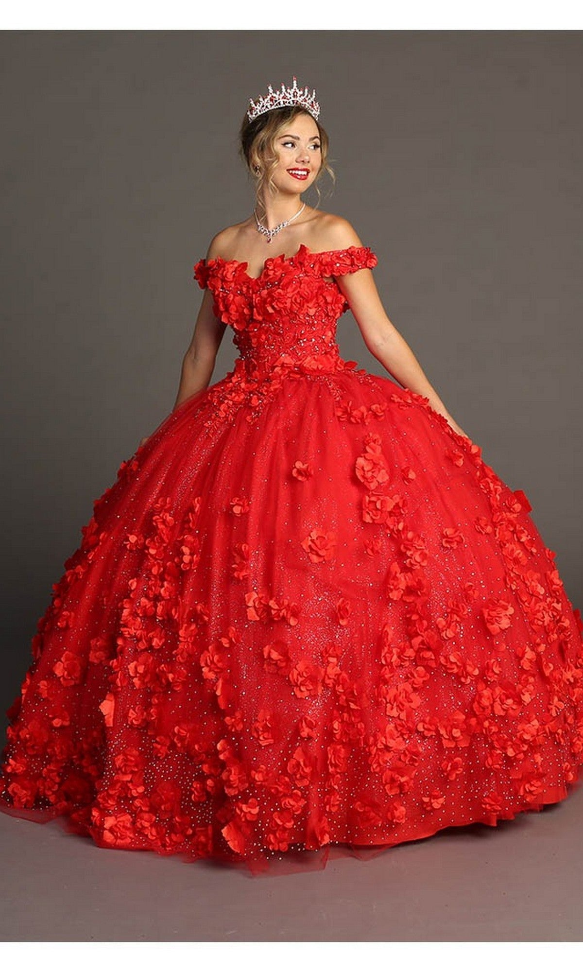 A woman stands against a plain gray background, wearing the exquisite Quinceanera Dress CHF3036 By Chicas in vibrant red—an off-shoulder tulle ballgown embellished with 3D floral accents—paired with a tiara and necklace, radiating elegance and charm.
