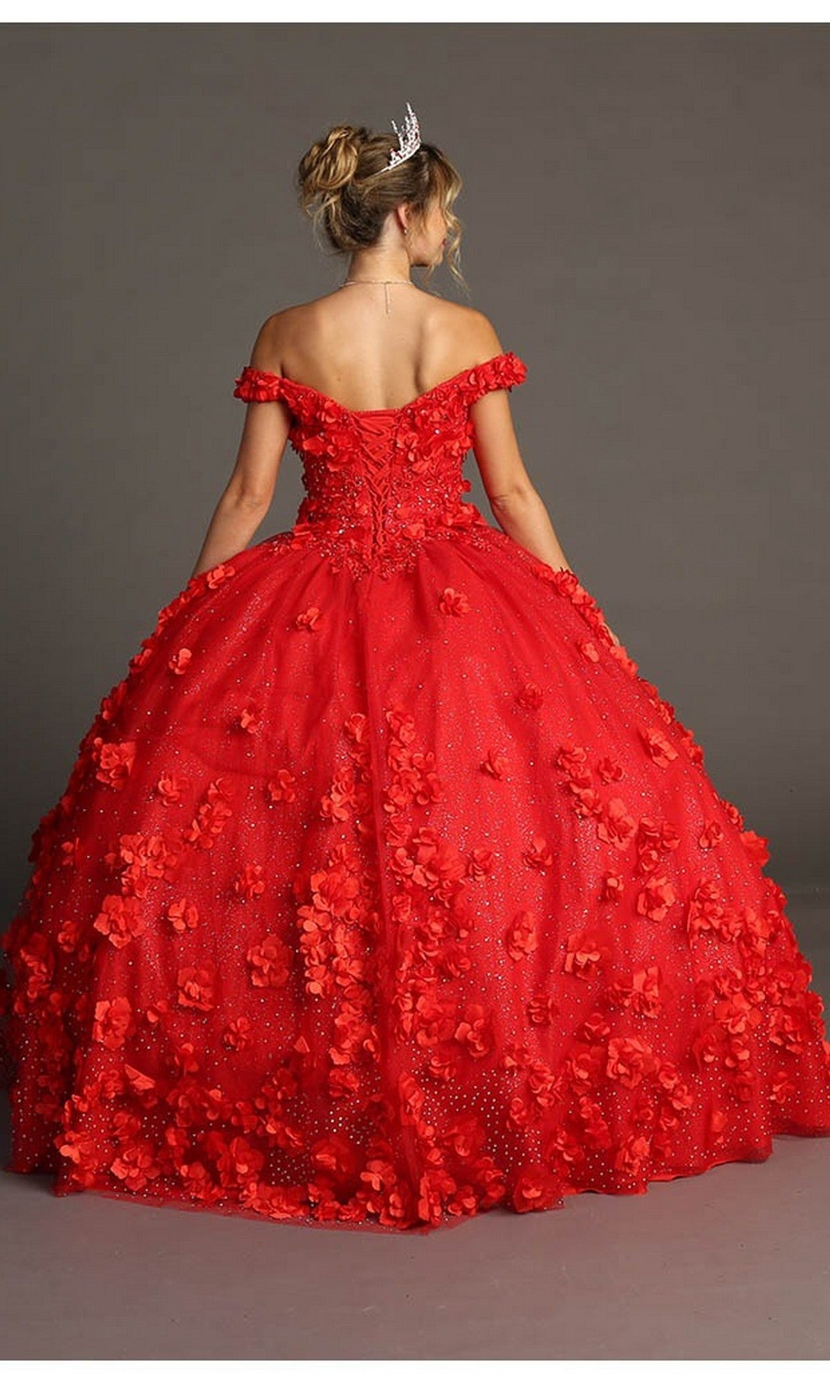 A woman wearing the Quinceanera Dress CHF3036 by Chicas—with off-shoulder styling, vibrant red color, and intricate 3D floral accents—pairs her look with a tiara as she stands against a plain gray background, facing away from the camera.