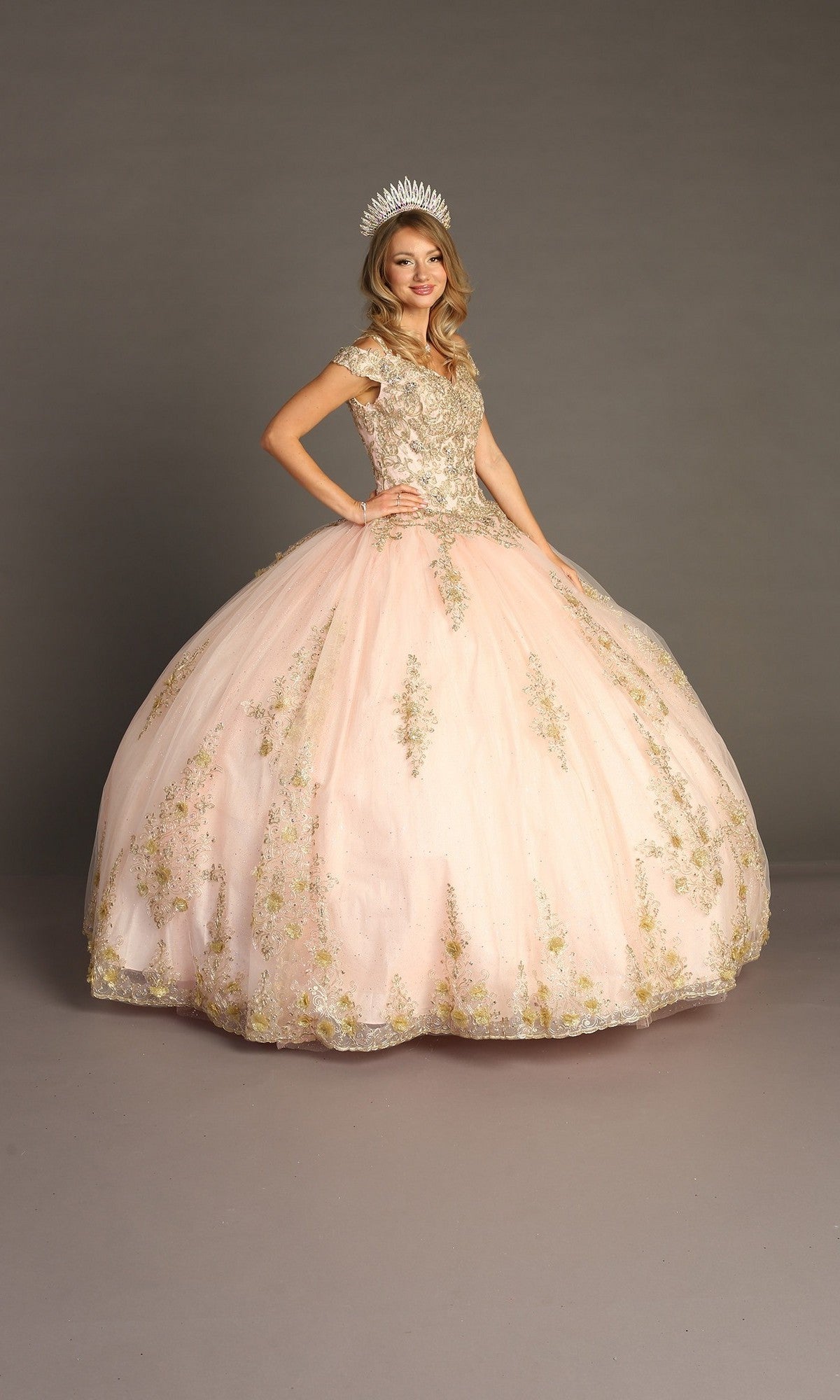 A young woman wears the Quinceanera Dress CHF3051 By Chicas in Blush/Gold, featuring ornate gold embroidery and an off-the-shoulder neckline; the elegant ballgown skirt flows gracefully as she stands against a plain gray background.
