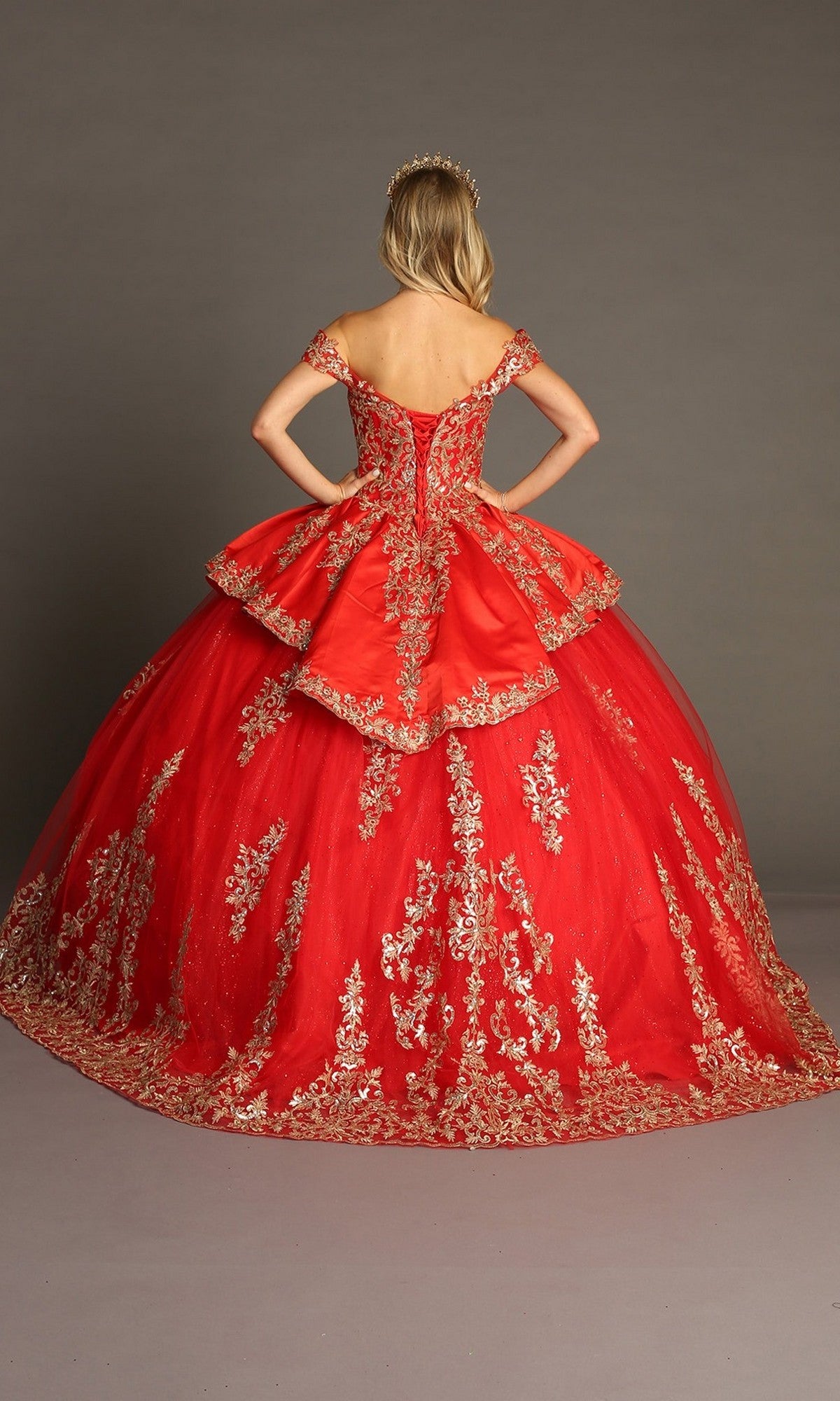 A woman stands with her back to the camera, hands on hips, wearing a gold tiara and showcasing the Quinceanera Dress CHF3052 By Chicas—a vibrant red gown adorned with gold embroidery—set against a plain studio background.