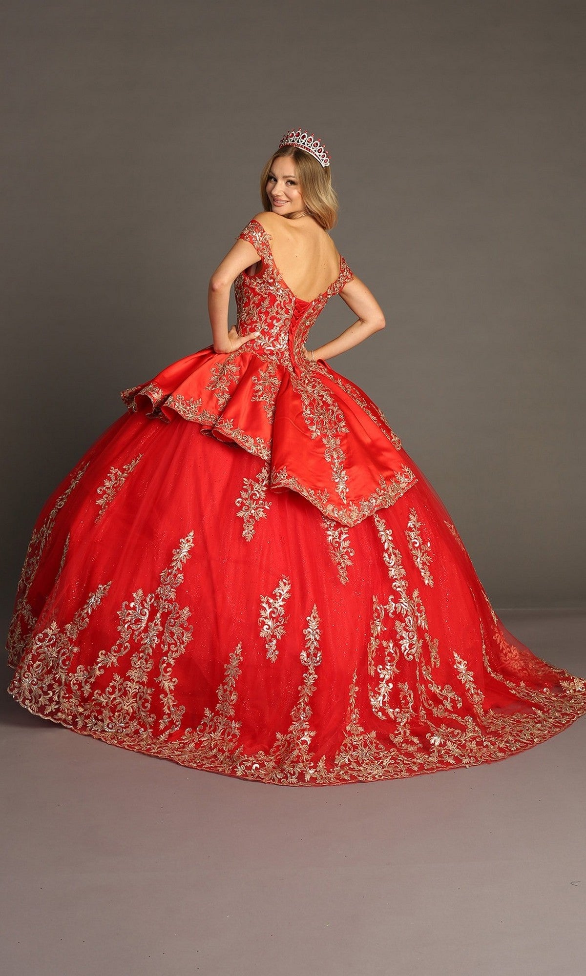 A woman wearing the Quinceanera Dress CHF3052 By Chicas—a red gown with gold embroidery and a tiara—stands on a gray backdrop, facing away and looking over her shoulder.