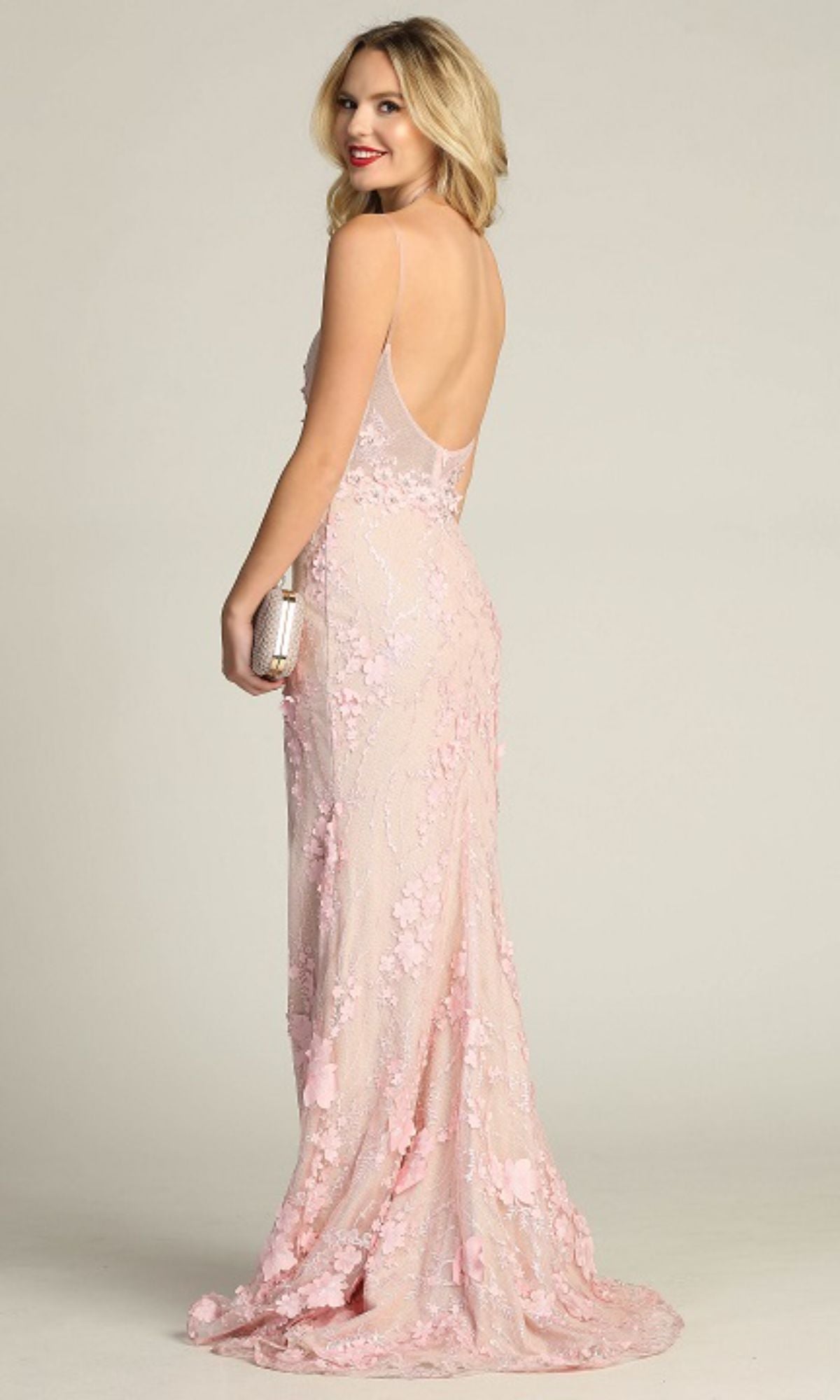 A woman wearing the Floral-Lace Long Prom Dress: Chicas CHF3058, a sleeveless, open-back light pink gown with floral detailing, stands holding a clutch and looks over her shoulder.