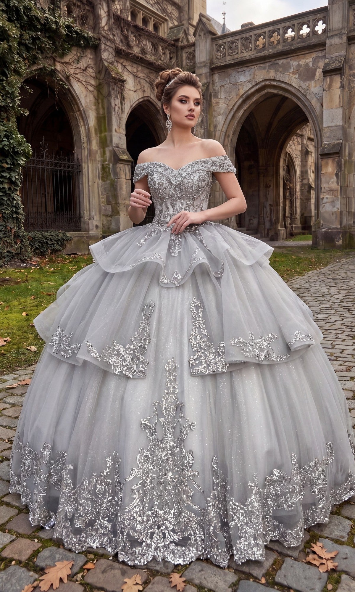 Wearing the Quinceanera Dress CHF3063 By Chicas in silver, a woman stands on a cobblestone path near a historic stone building with arches. The off-the-shoulder gown features ornate lace details and sparkly embellishments.