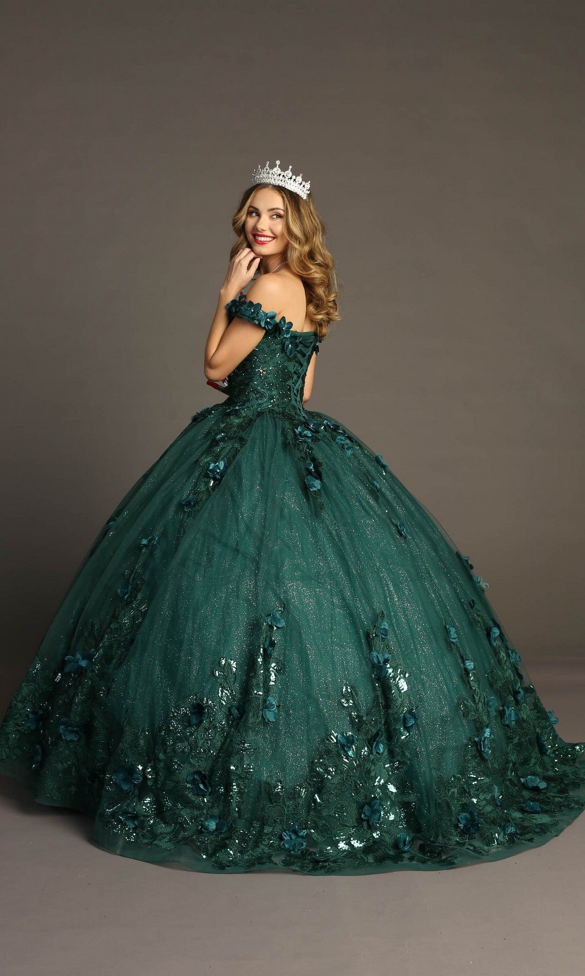A woman wearing the Quinceanera Dress CHF3069 By Chicas—a green off-the-shoulder ballgown embellished with 3D floral appliques—stands against a gray background, smiling and crowned.
