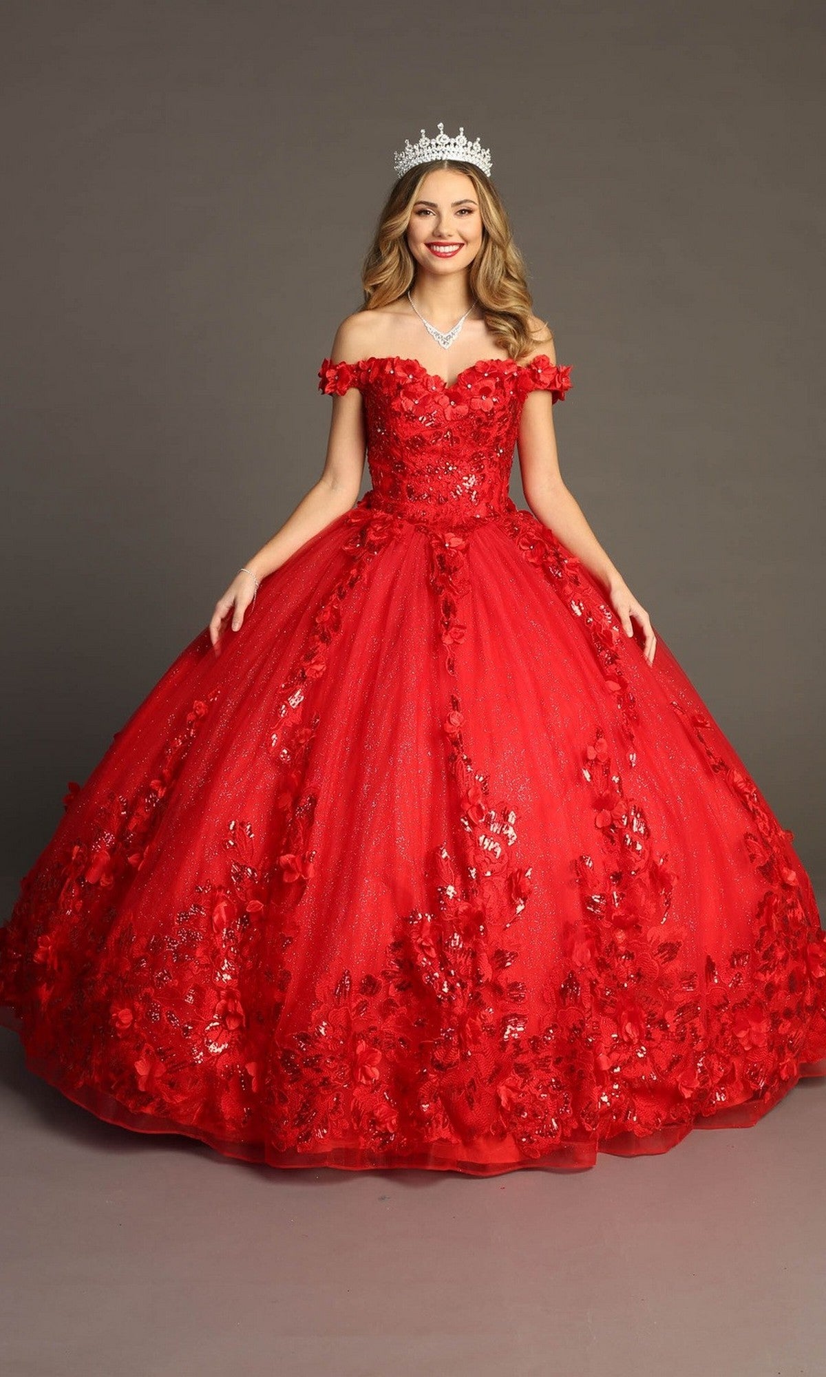 A woman wearing the Quinceanera Dress CHF3069 by Chicas in red, featuring an off-the-shoulder design with intricate 3D floral appliques and a silver crown, stands against a plain background, smiling.