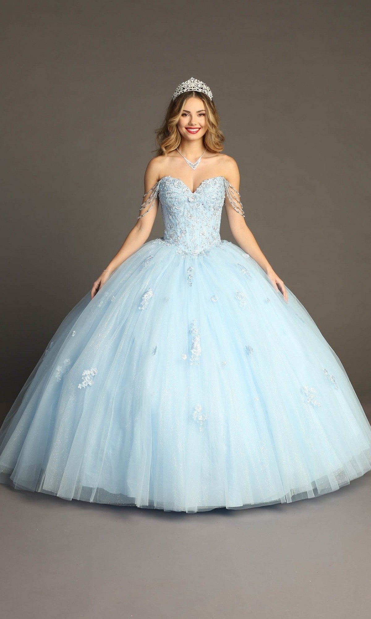 A woman poses against a plain studio backdrop wearing the Quinceanera Dress CHF3071 by Chicas—a light blue glitter-tulle ballgown featuring off-the-shoulder sleeves and a beaded bodice—styled with a tiara and necklace.
