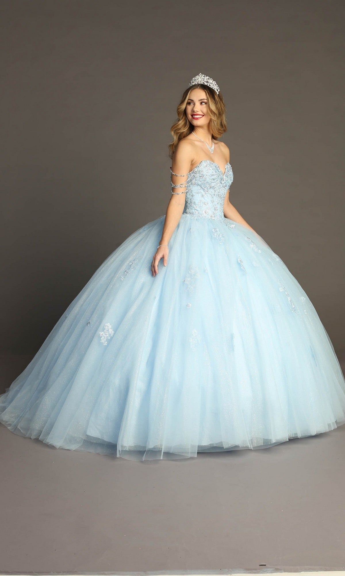 A woman stands and smiles against a plain gray background, wearing the Quinceanera Dress CHF3071 By Chicas—a light blue glitter-tulle ballgown with a beaded bodice and a silver tiara.