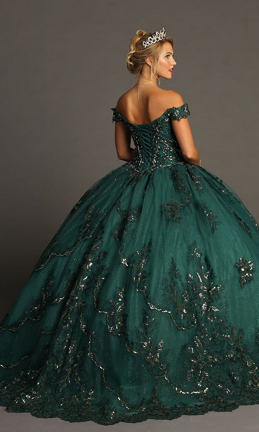A woman wearing the Quinceanera Dress CHF3072 by Chicas—an elegant dark green, off-the-shoulder gown featuring glittering metallic embroidery and a flowing ballgown skirt—stands against a neutral background, crowned with a tiara.