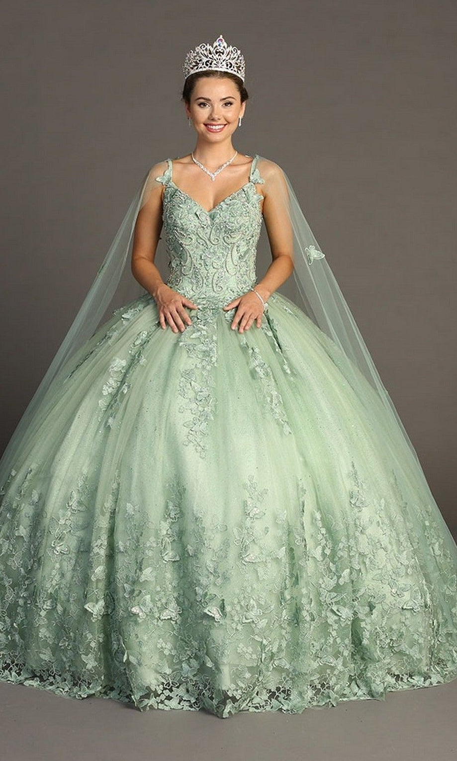 A woman wearing the Quinceanera Dress CHF3077 By Chicas—a mint green glitter tulle ballgown with lace detailing—along with a tiara and a necklace, stands against a plain gray background, smiling at the camera.