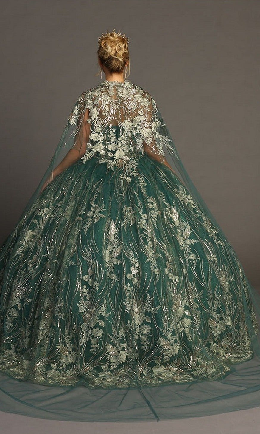 A woman stands facing away against a plain backdrop, wearing the Quinceanera Dress CHF3079 By Chicasโa strapless green ballgown adorned with metallic floral embroidery and paired with a sheer, embroidered cape.