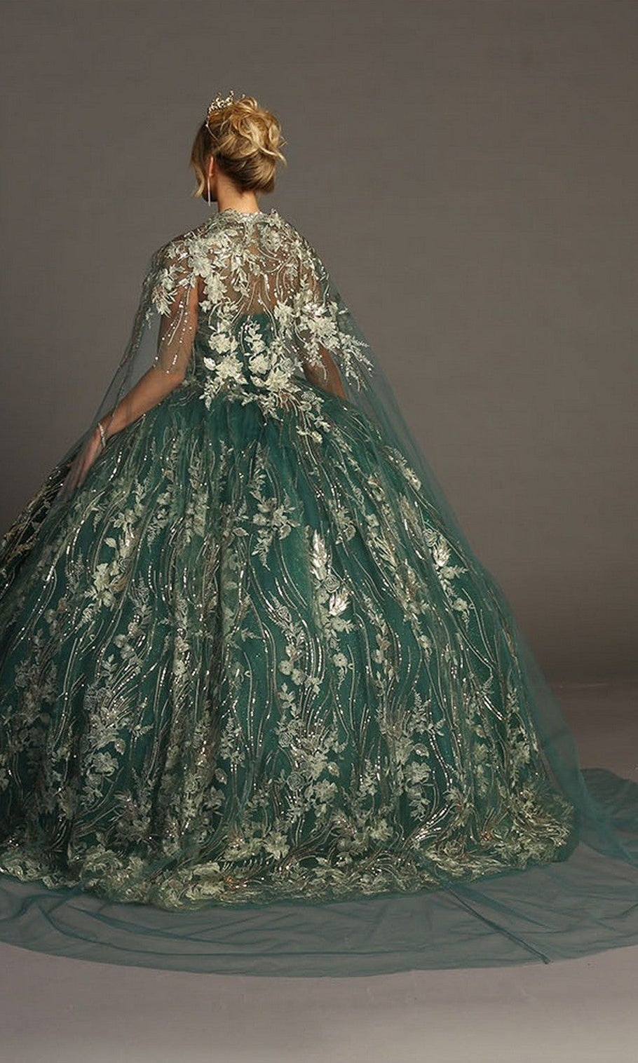 A woman in the Quinceanera Dress CHF3079 by Chicasโa strapless green ballgown featuring metallic floral embroidery and an embroidered capeโstands facing away, her hair styled in an updo adorned with a tiara.