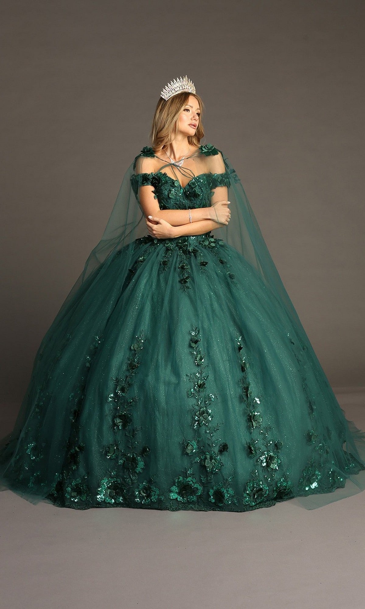 A woman wearing the Quinceanera Dress CHF3082 By Chicas, a dark green gown adorned with 3D floral appliques and paired with a tiara, stands against a plain background with her arms crossed.