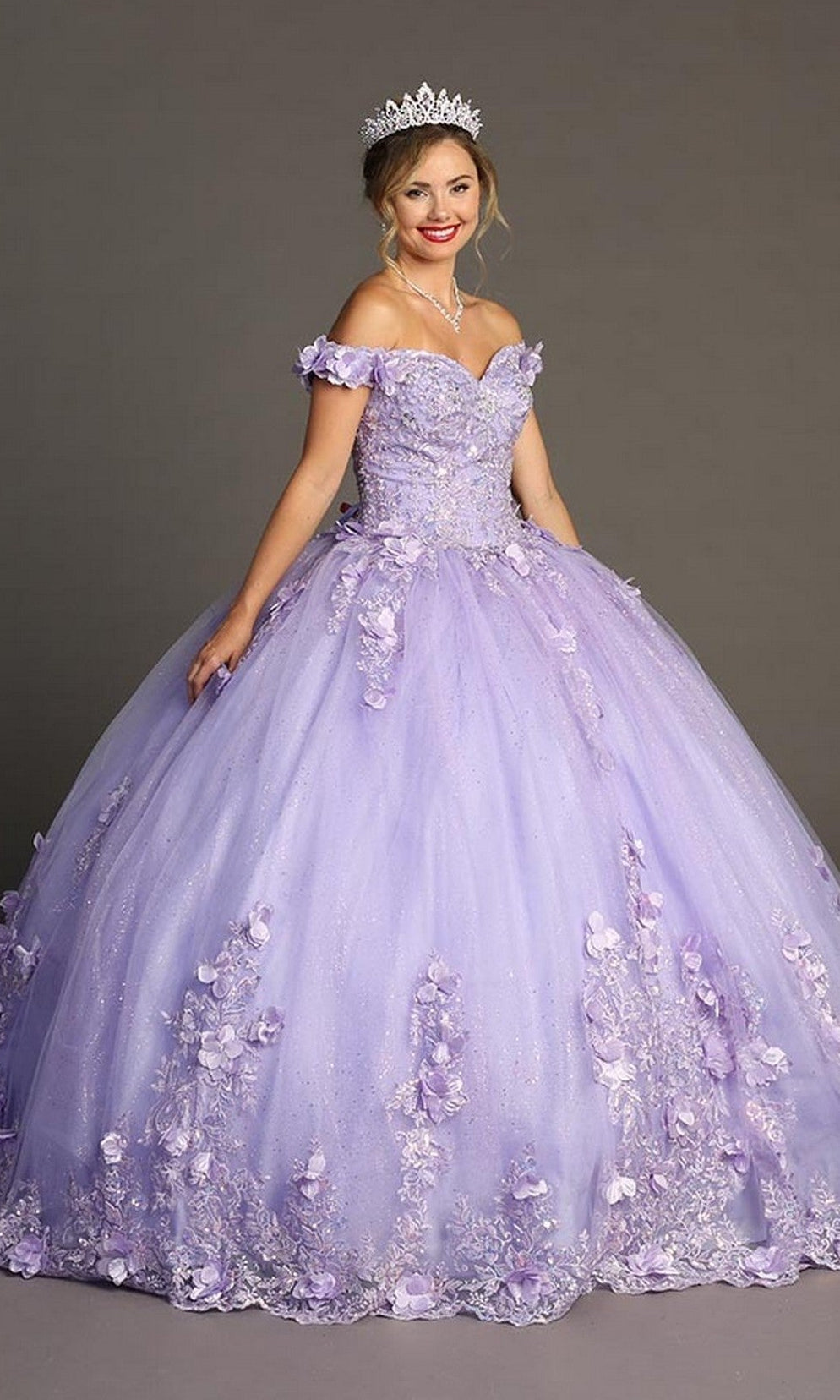 A young woman wearing the Quinceanera Dress CHF3085 By Chicas—a lavender, off-the-shoulder gown adorned with floral appliqués and complemented by a tiara—stands against a plain gray background.