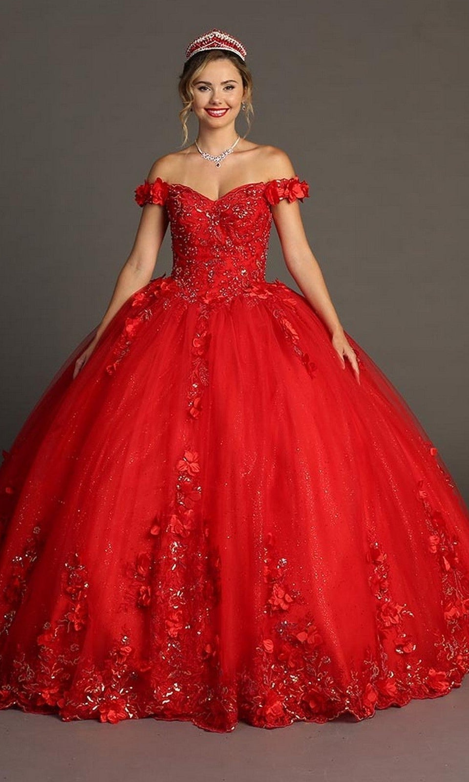 A woman wearing the red Quinceanera Dress CHF3085 by Chicas, featuring floral appliques and an off-the-shoulder design, paired with a sparkling tiara and necklace, stands against a plain gray background.