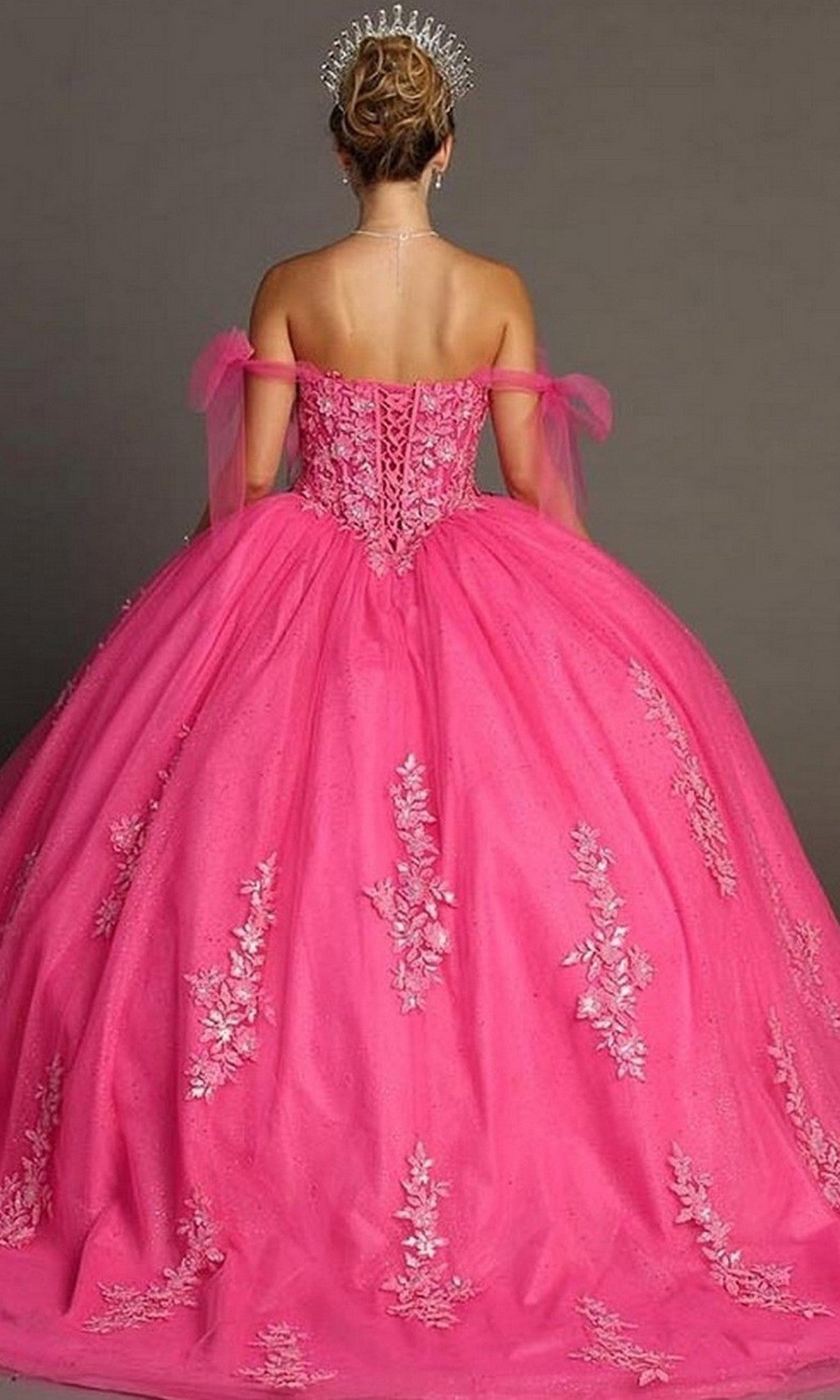 A woman is shown from the back against a plain background, highlighting the elegance of her Quinceanera Dress CHF3086 By Chicas—a bright pink off-the-shoulder ball gown adorned with floral embellishments and paired with a tiara, reminiscent of a classic quinceañera style.