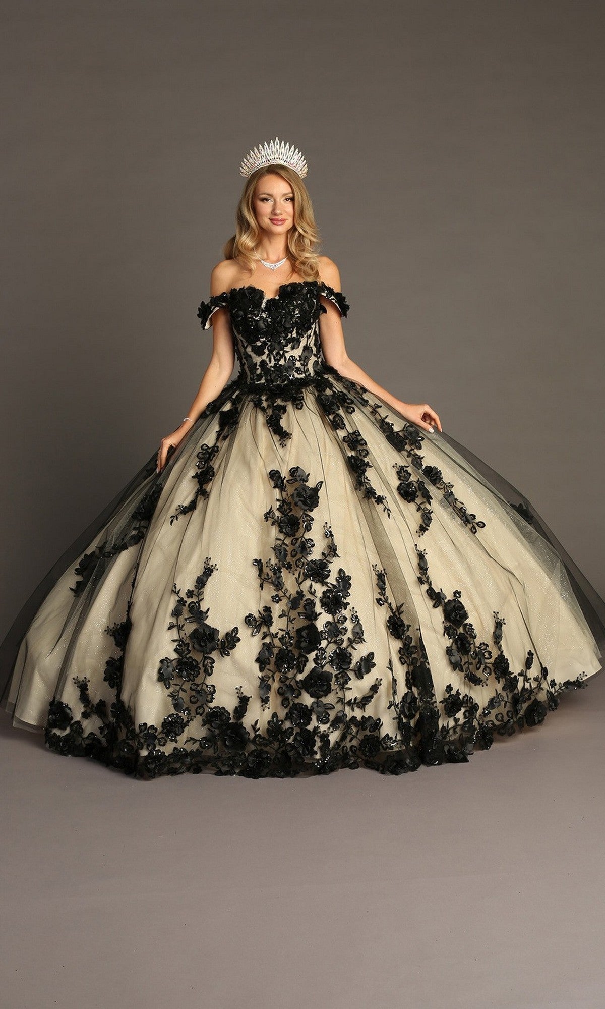 Woman wearing the Quinceanera Dress CHF3088 By Chicas in black/nude, an off-the-shoulder tulle ballgown featuring black floral appliqué and a full skirt, accessorized with a tiara and standing against a plain background.