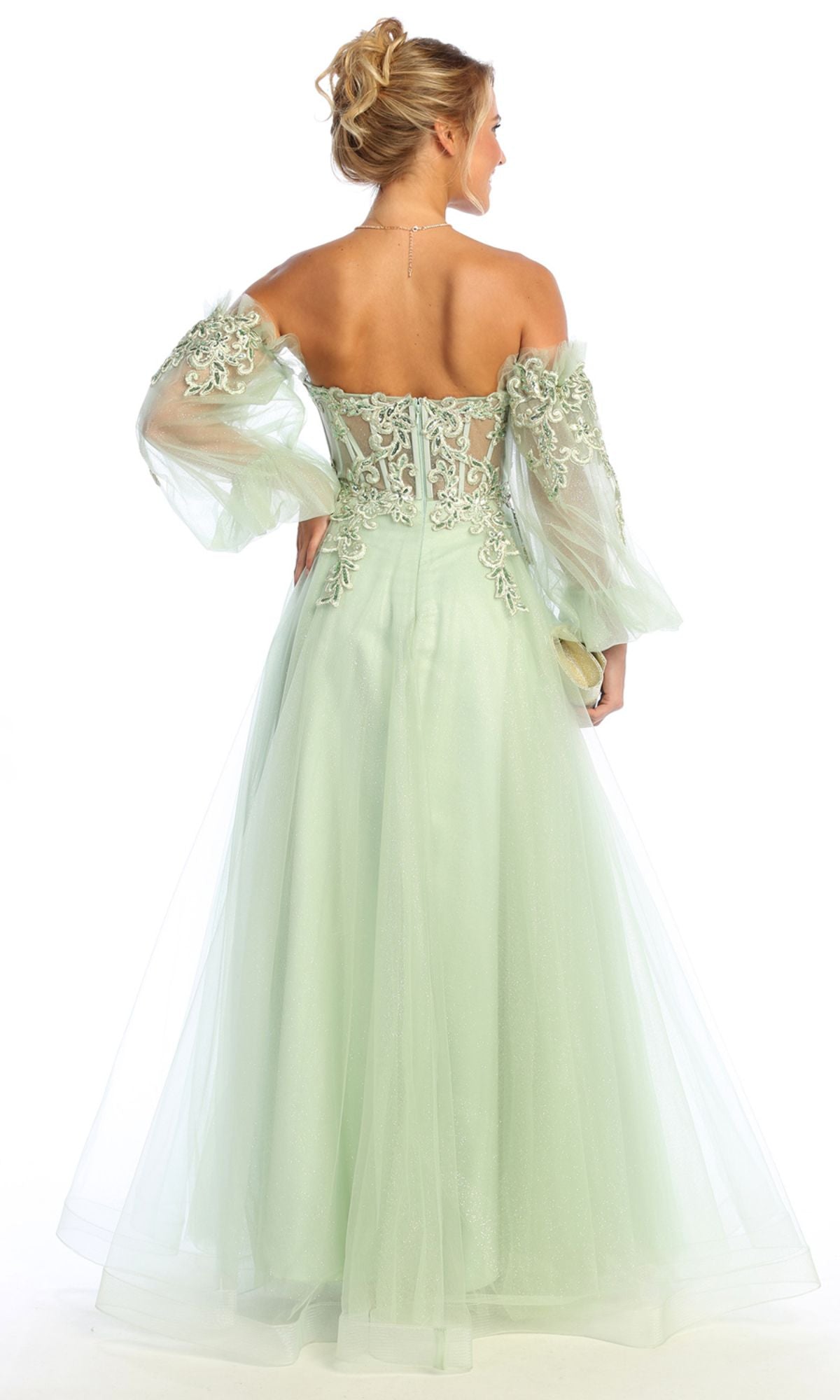 A woman in the Chicas CHF3093 long sleeve A-line prom dress with lace-up back stands facing away, wearing a light green sheer embroidered off-the-shoulder gown against a white background.