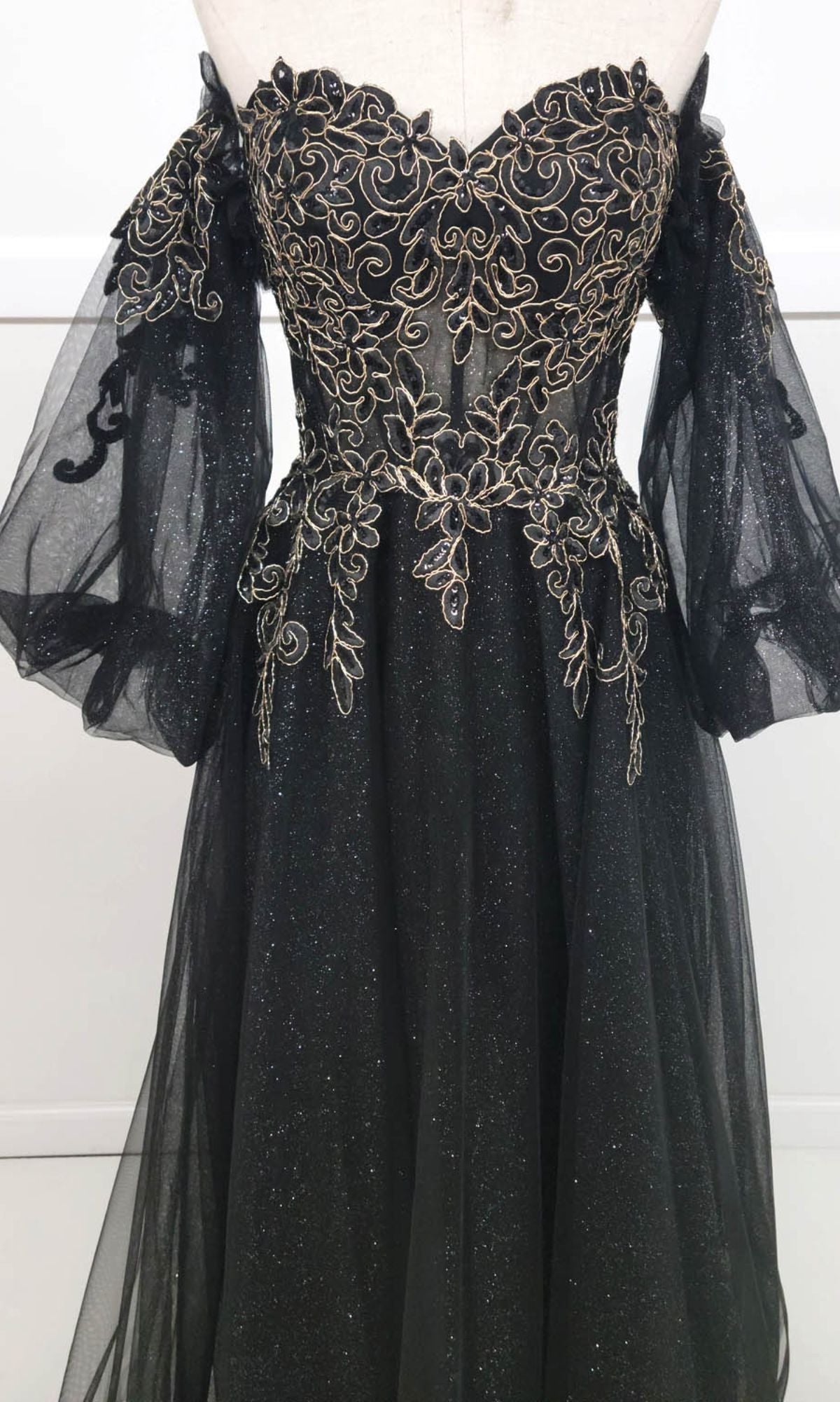 The Chicas CHF3093 Long Sleeve Long A-Line Prom Dress is a black gown with gold floral embroidery, sheer billowing sleeves, and an off-the-shoulder design, shown on a dress form against a plain background.