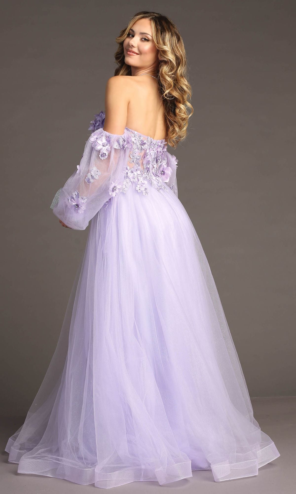 A woman with wavy blonde hair models the Chicas CHF3096 Long Sleeve Long Prom Ball Gown in lavender, featuring floral appliqués and sheer sleeves, as she poses against a plain background.