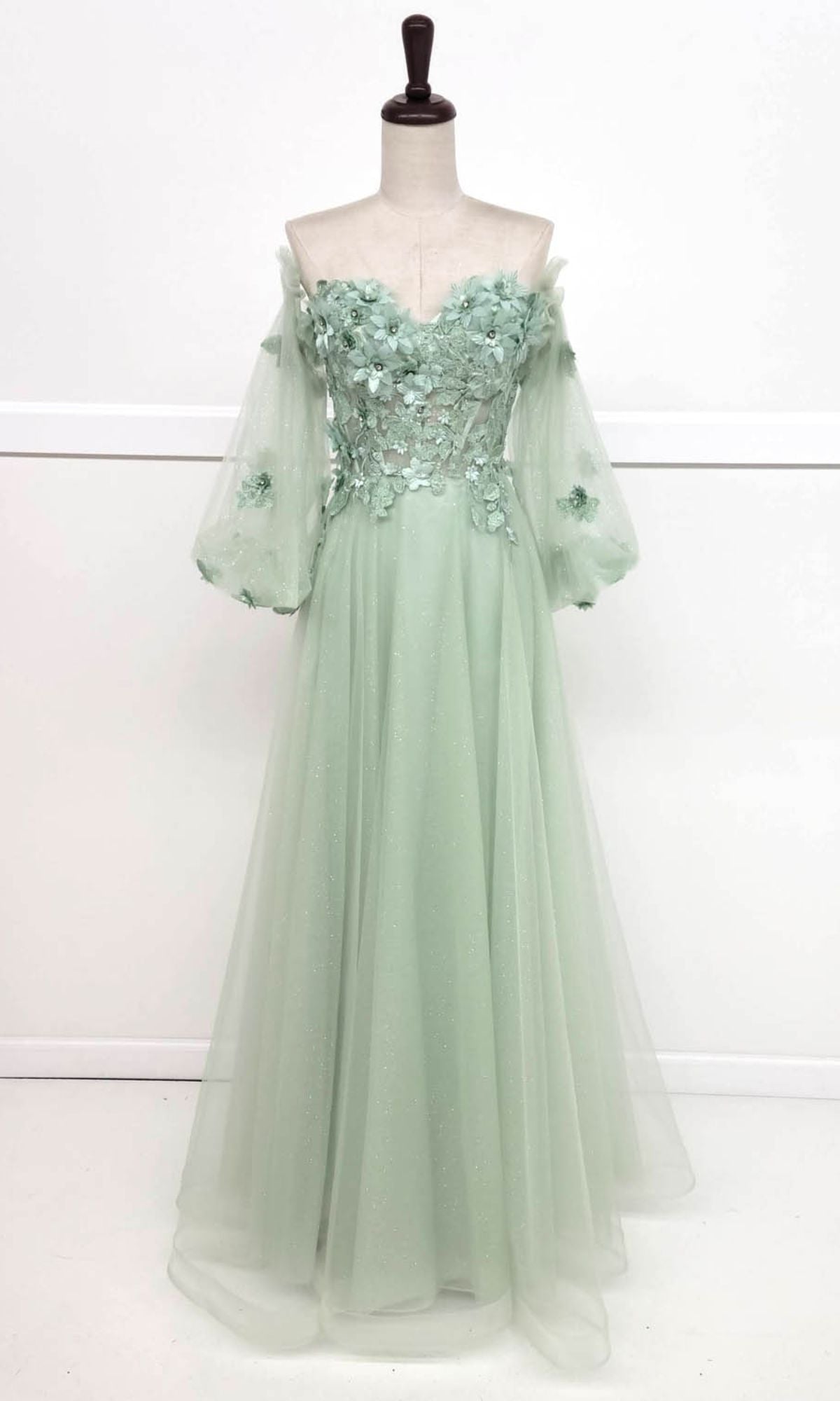 The Chicas CHF3096 Long Sleeve Long Prom Ball Gown in sage green features an off-the-shoulder design, sheer puff sleeves, a floral appliqué bodice, and a flowing floor-length tulle skirt displayed on a dress form.